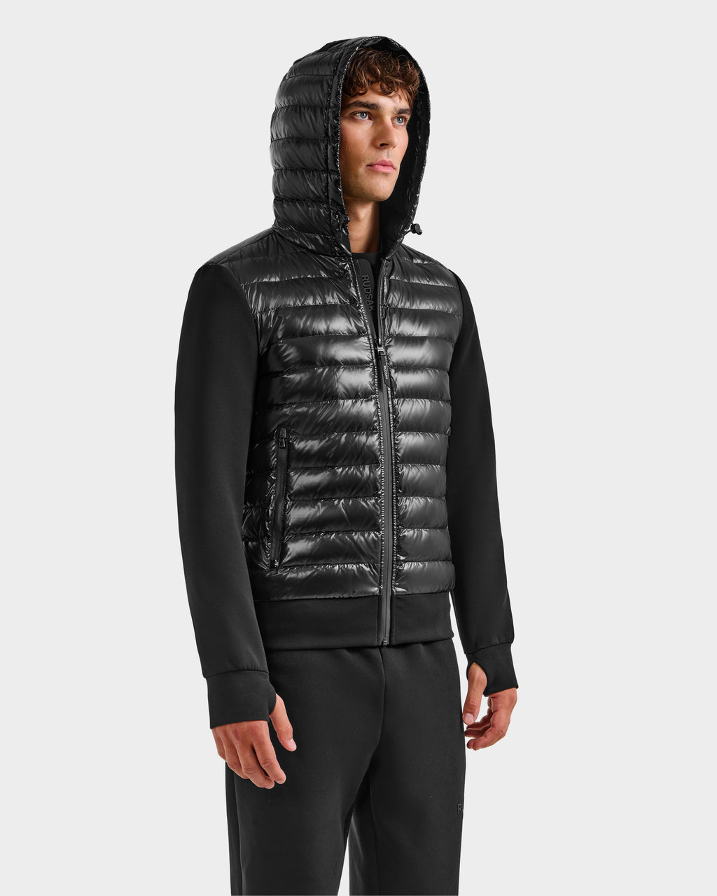 A man stands slightly to the side against a plain light background wearing the Rudsak DEE S - 6125684 Black men’s lightweight down jacket, featuring an RDS-certified duck down fill, shiny quilted front, and matte black sleeves.