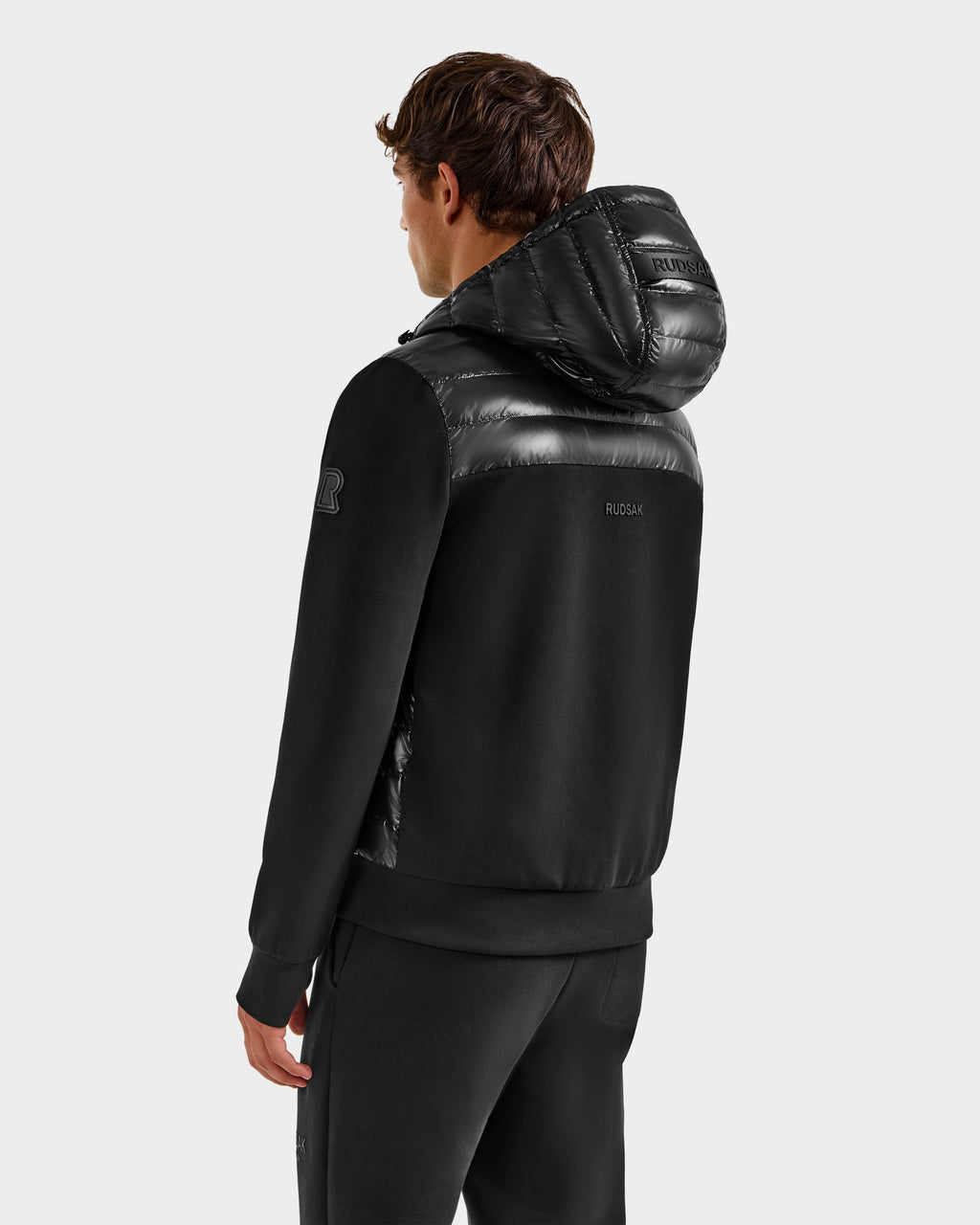 A man facing away from the camera wears the Rudsak DEE S - 6125684 Black men’s lightweight down jacket with shiny quilted hood and shoulders, paired with black pants against a plain light gray background—ideal for chilly urban days.