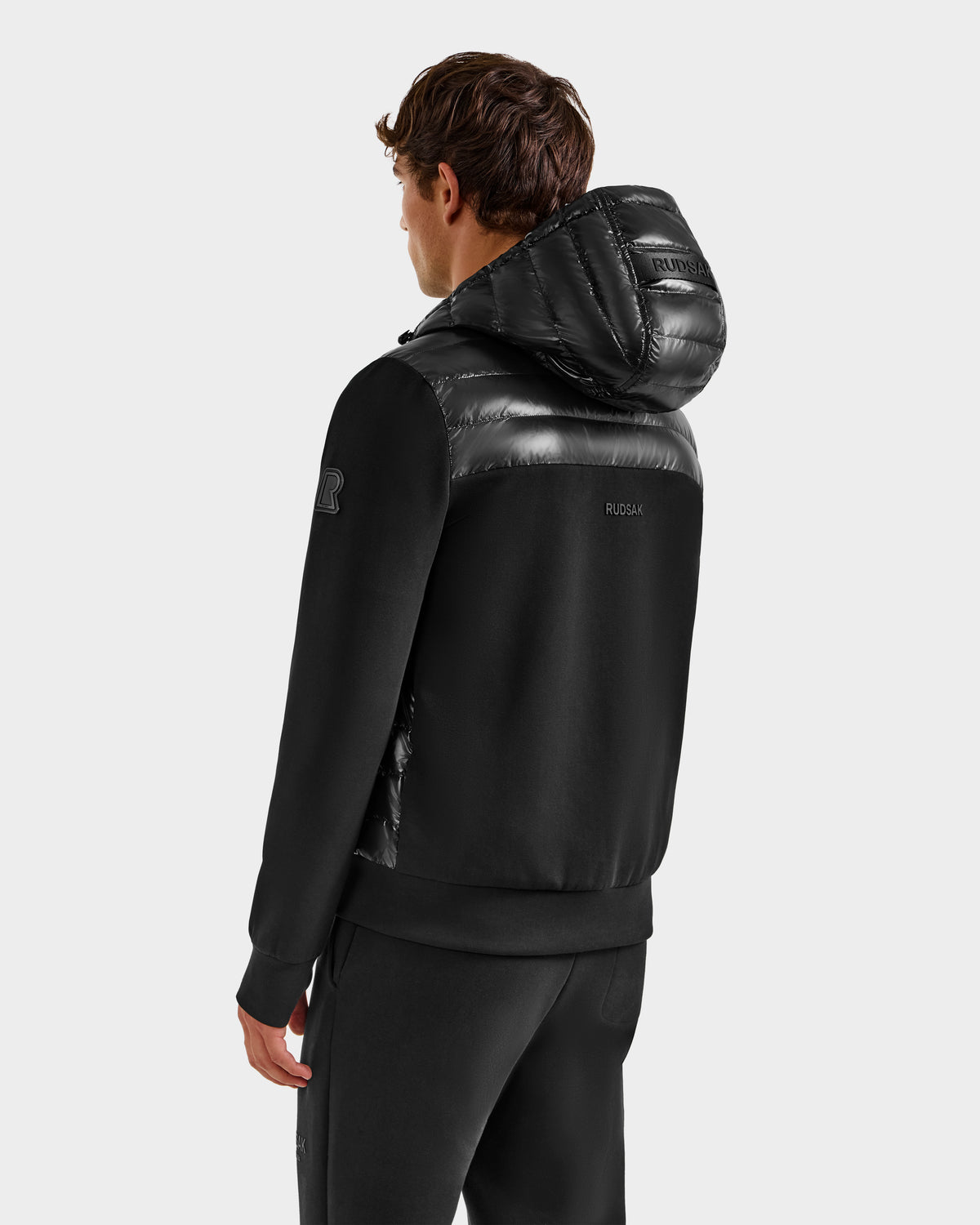 A man facing away from the camera wears the Rudsak DEE S - 6125684 Black men’s lightweight down jacket with shiny quilted hood and shoulders, paired with black pants against a plain light gray background—ideal for chilly urban days.