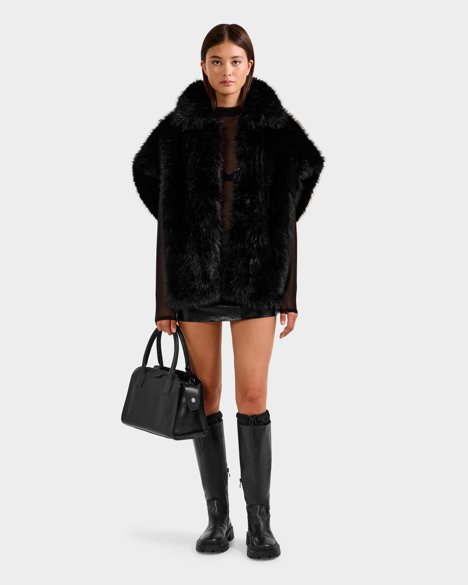 A woman stands facing forward in an oversized silhouette, wearing the Rudsak DELFINA - 8525662 Black faux fur cape. She pairs it with a short black skirt, knee-high boots, and holds a black handbag against a light gray background.