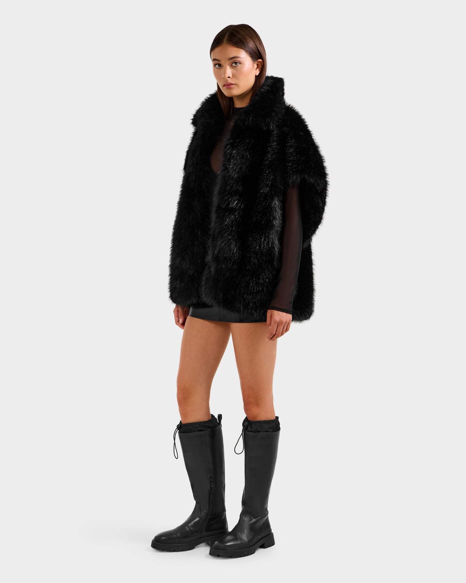 A woman wears the Rudsak DELFINA - 8525662 Black faux fur vest over a sheer black top, short skirt, and tall boots—an effortlessly chic winter look against a plain white background.