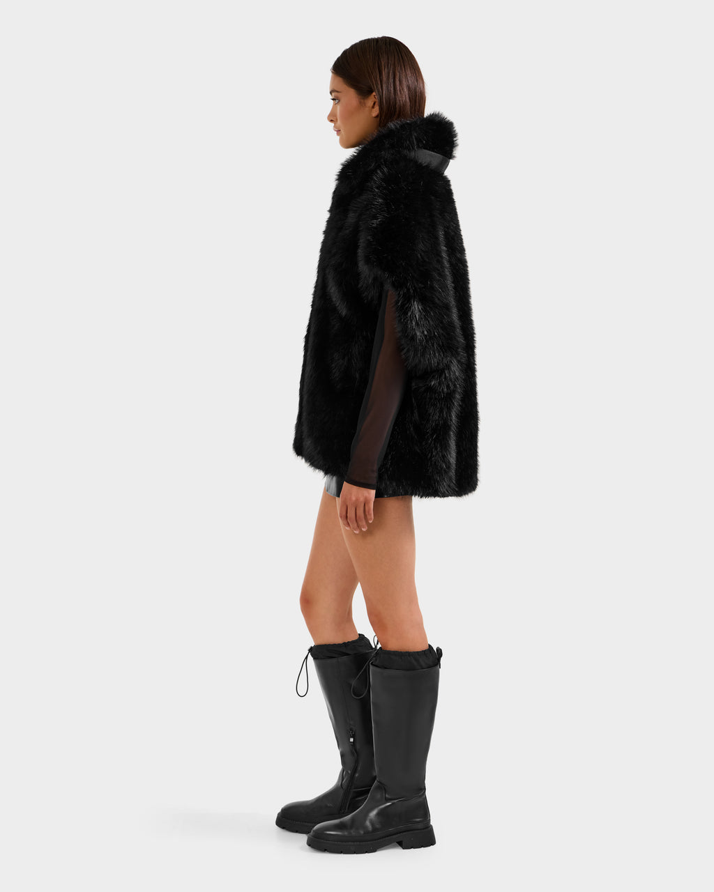 A woman in profile wears the Rudsak DELFINA - 8525662 Black faux fur coat with an oversized silhouette, styled with knee-high boots and a black mini skirt, set against a plain light gray background.