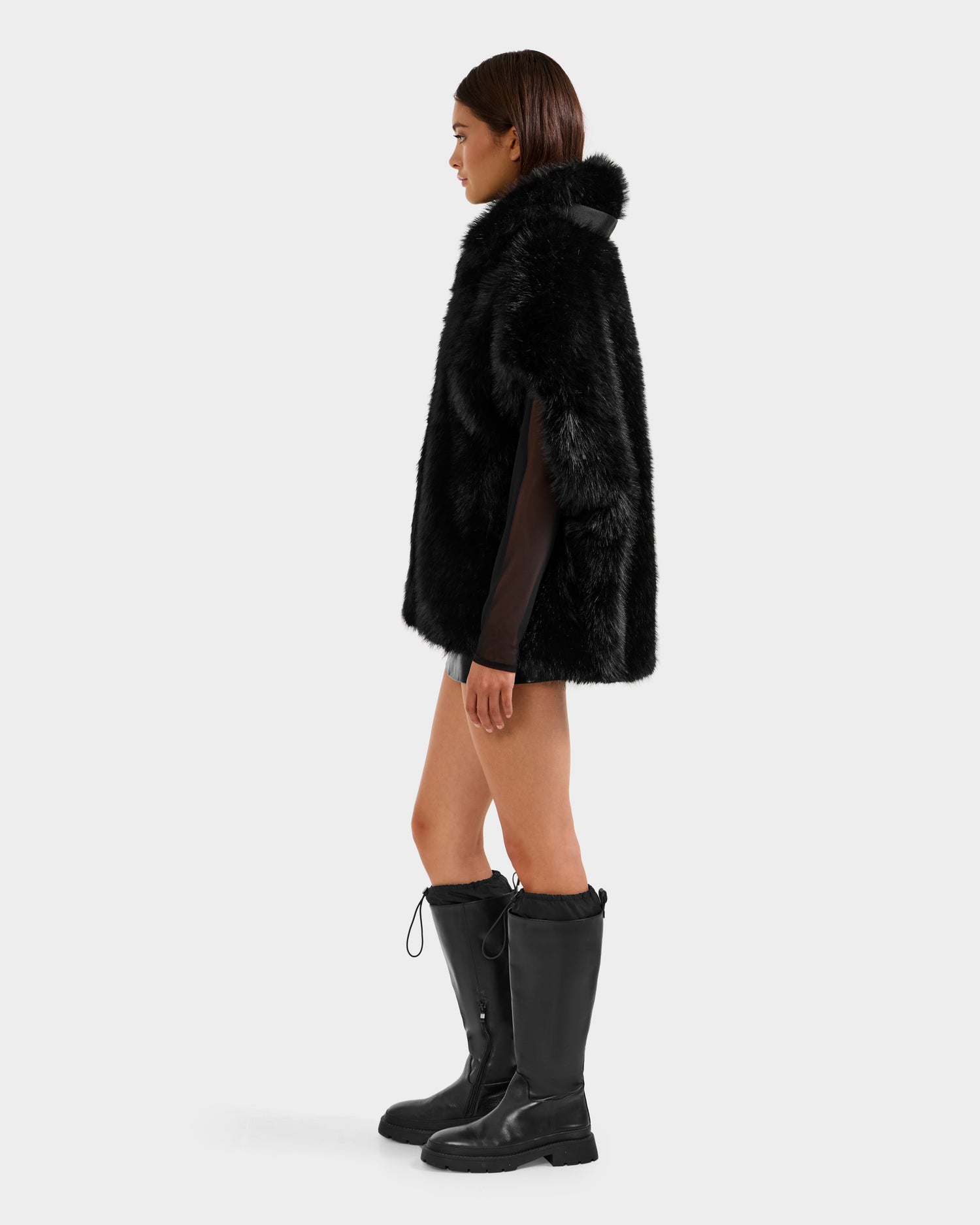 A woman in profile wears the Rudsak DELFINA - 8525662 Black faux fur coat with an oversized silhouette, styled with knee-high boots and a black mini skirt, set against a plain light gray background.