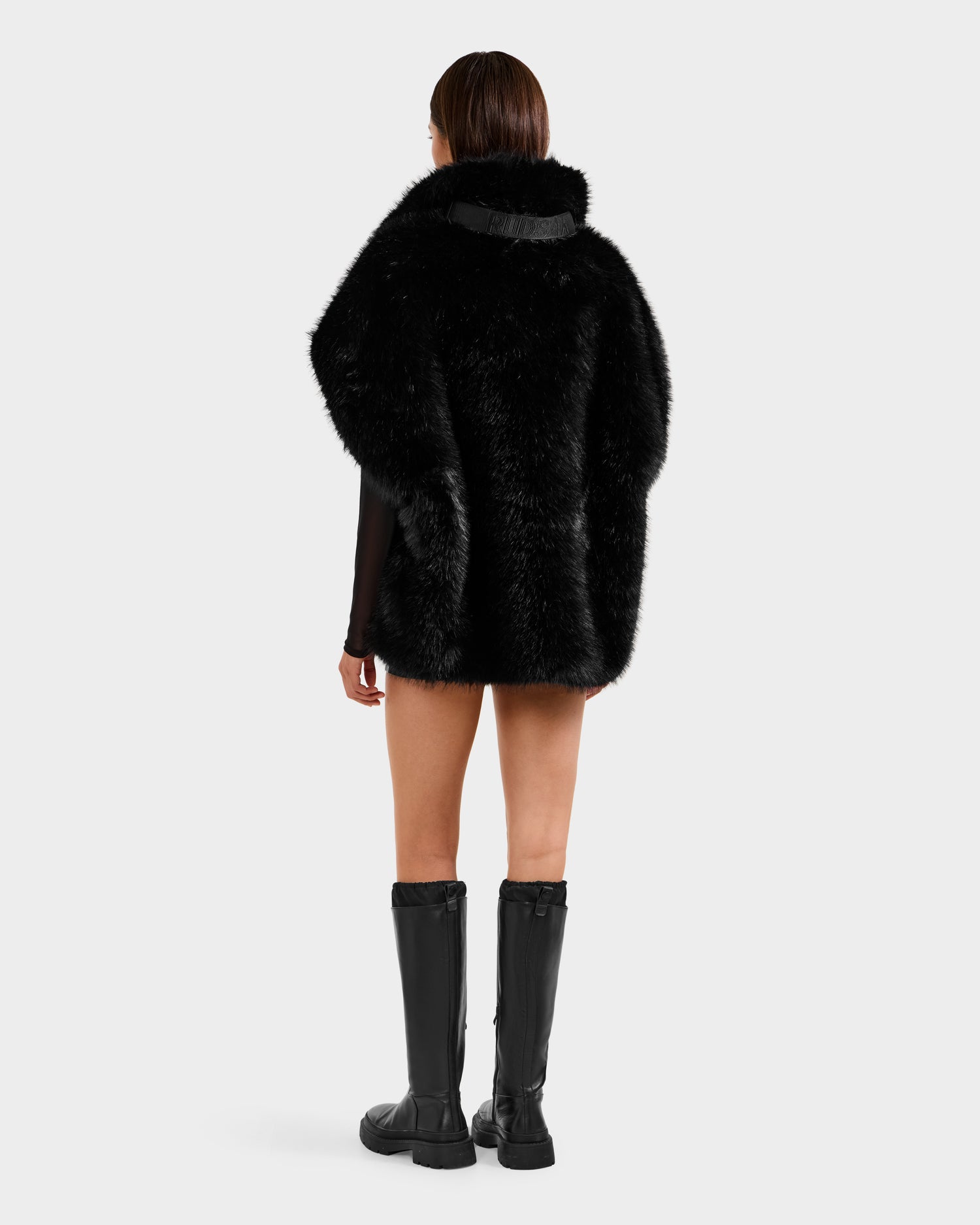 A person with straight brown hair faces away, wearing Rudsak's DELFINA - 8525662 Black faux fur coat with an oversized silhouette and black knee-high boots. The background is plain and light, offering chic winter wardrobe inspiration.