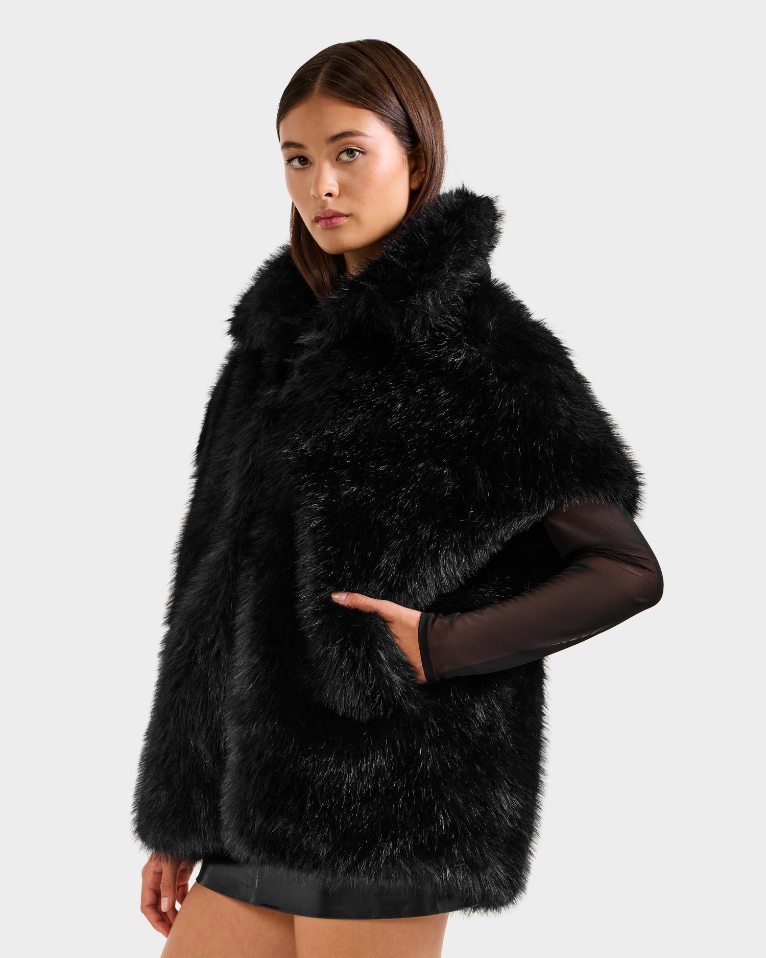 A woman with straight brown hair models the Rudsak DELFINA - 8525662 Black faux fur cape with a high collar and mesh sleeve, standing against a light background—an elegant option for transitional style.