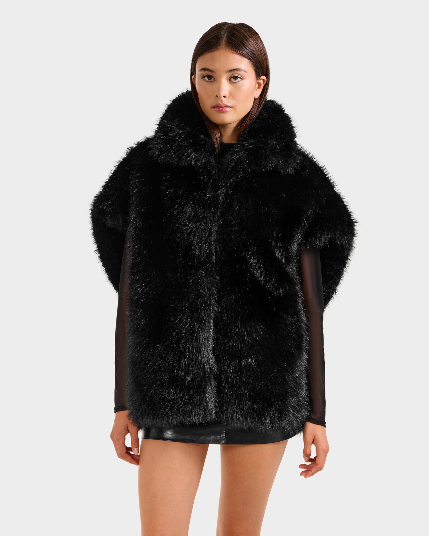 A woman with straight brown hair and a neutral expression models the Rudsak DELFINA - 8525662 Black faux fur coat, featuring an oversized silhouette and high collar, against a plain light grey background.
