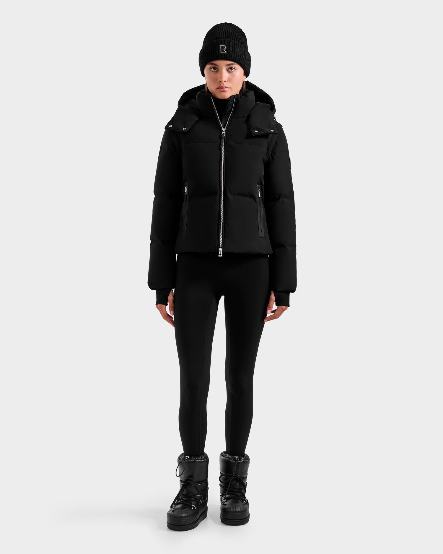 A person stands facing forward, wearing the Rudsak DELILAH M - L125538 Black women’s down puffer with removable sleeves, paired with black leggings, a black beanie, and black boots against a plain light gray background.