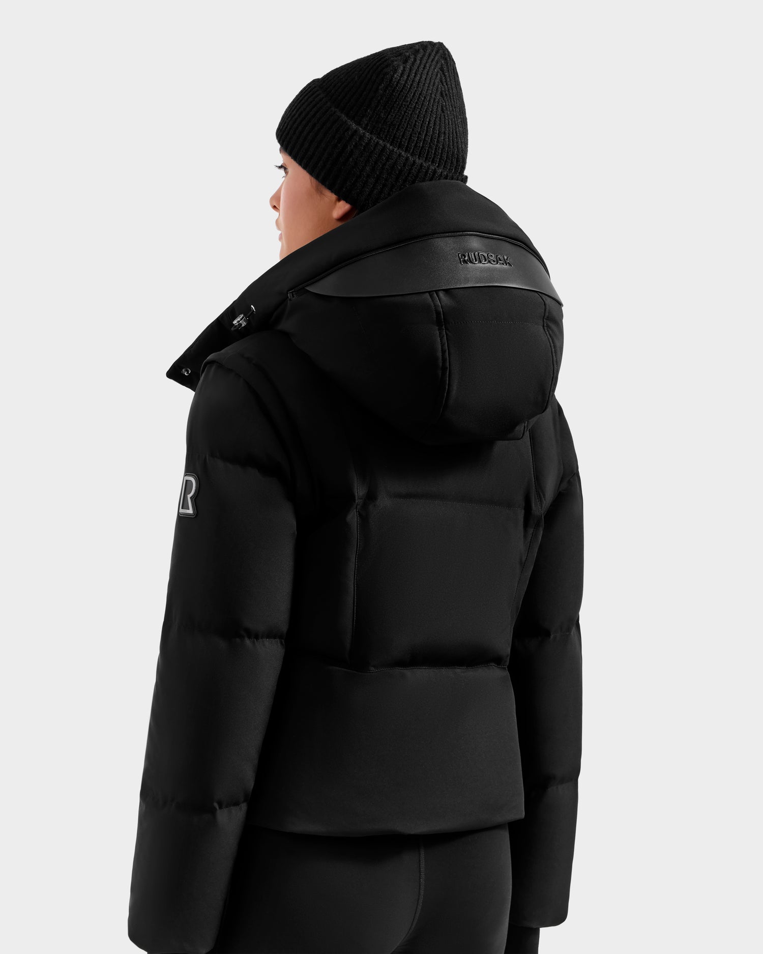 A person is seen from the back and side against a plain light background wearing Rudsak’s DELILAH M - L125538 Black women’s down puffer jacket with hood, visible logo on the sleeve and collar, black knit beanie, and black pants.