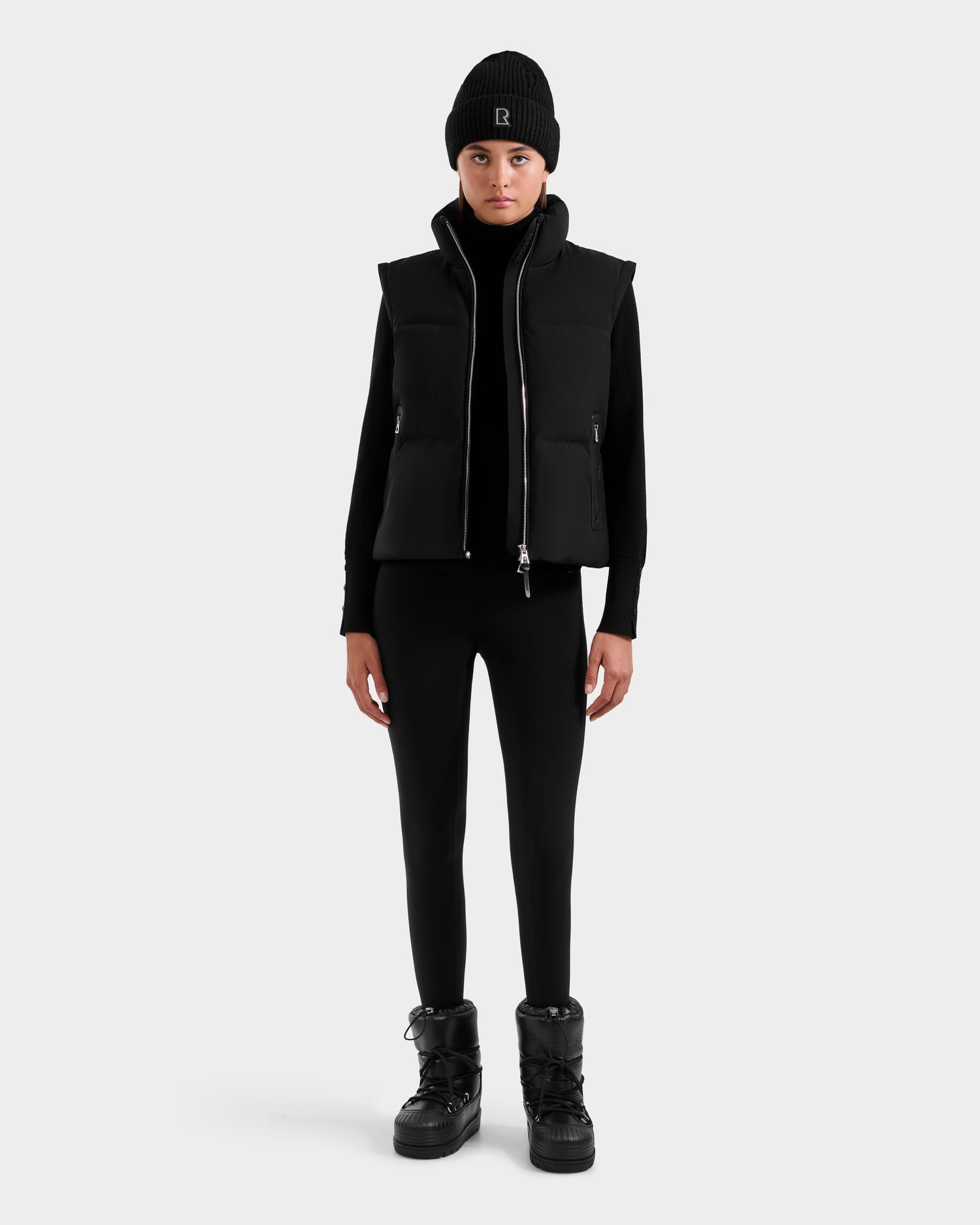 A person stands facing forward wearing the Rudsak DELILAH M - L125538 Black women’s down puffer with removable sleeves, a black long-sleeve top, leggings, and lace-up boots against a plain light gray background.
