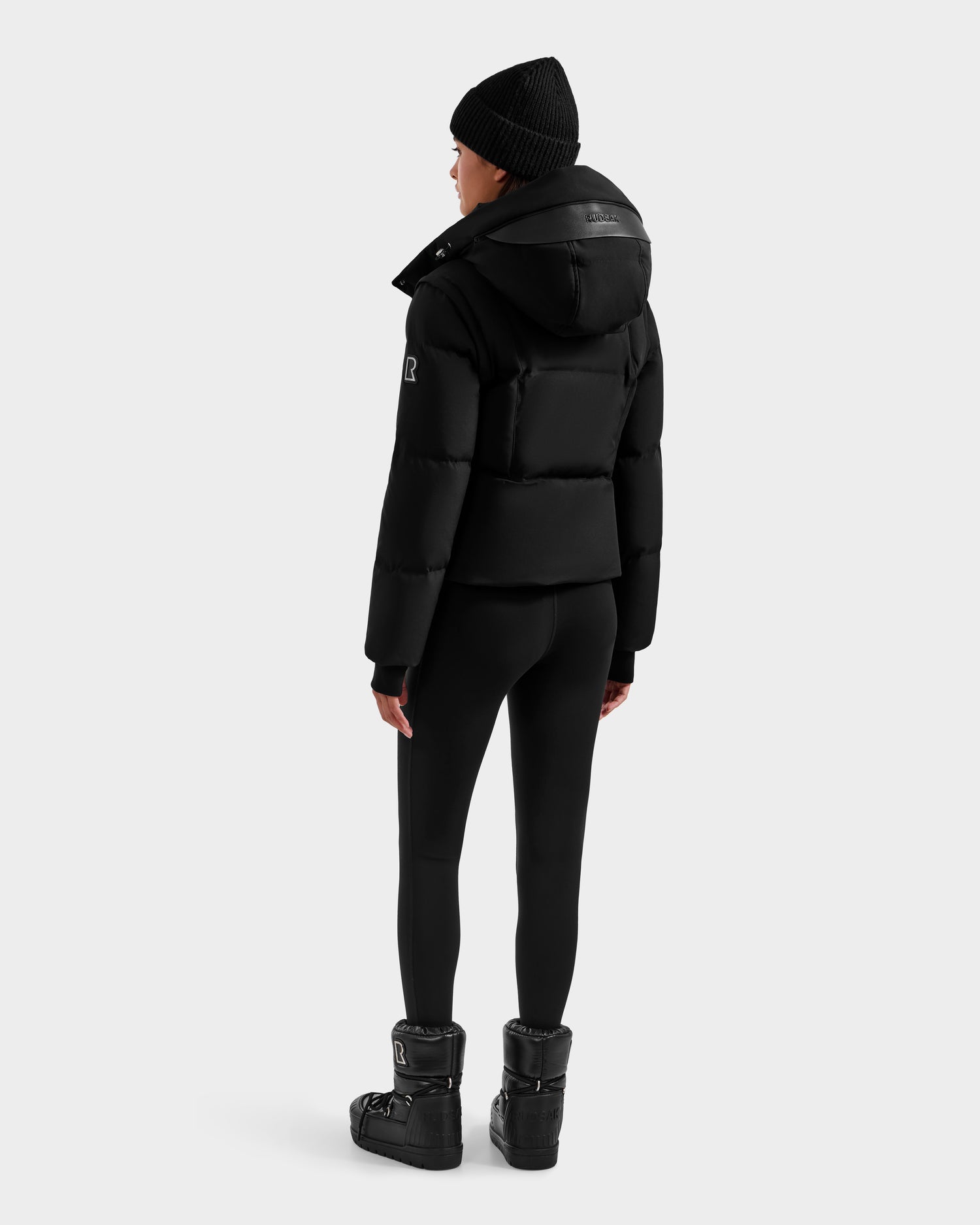 A person wearing a Rudsak DELILAH M - L125538 Black women’s down puffer with removable sleeves, black leggings, winter boots, and a black beanie stands facing away from the camera against a plain light gray background.