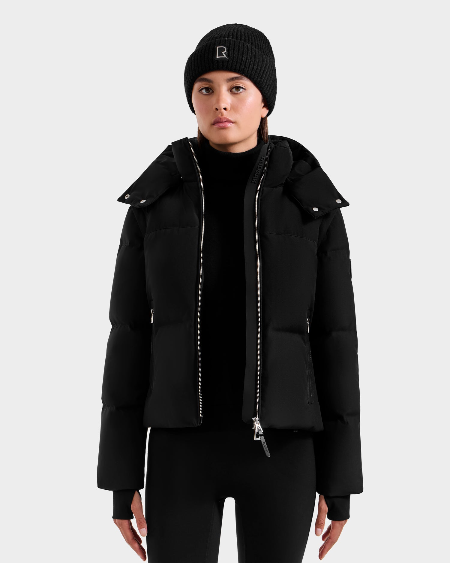 A person faces forward in a neutral pose, wearing the Rudsak DELILAH M - L125538 Black women’s down puffer with removable sleeves, paired with a black knit beanie and black pants, set against a plain light gray background.