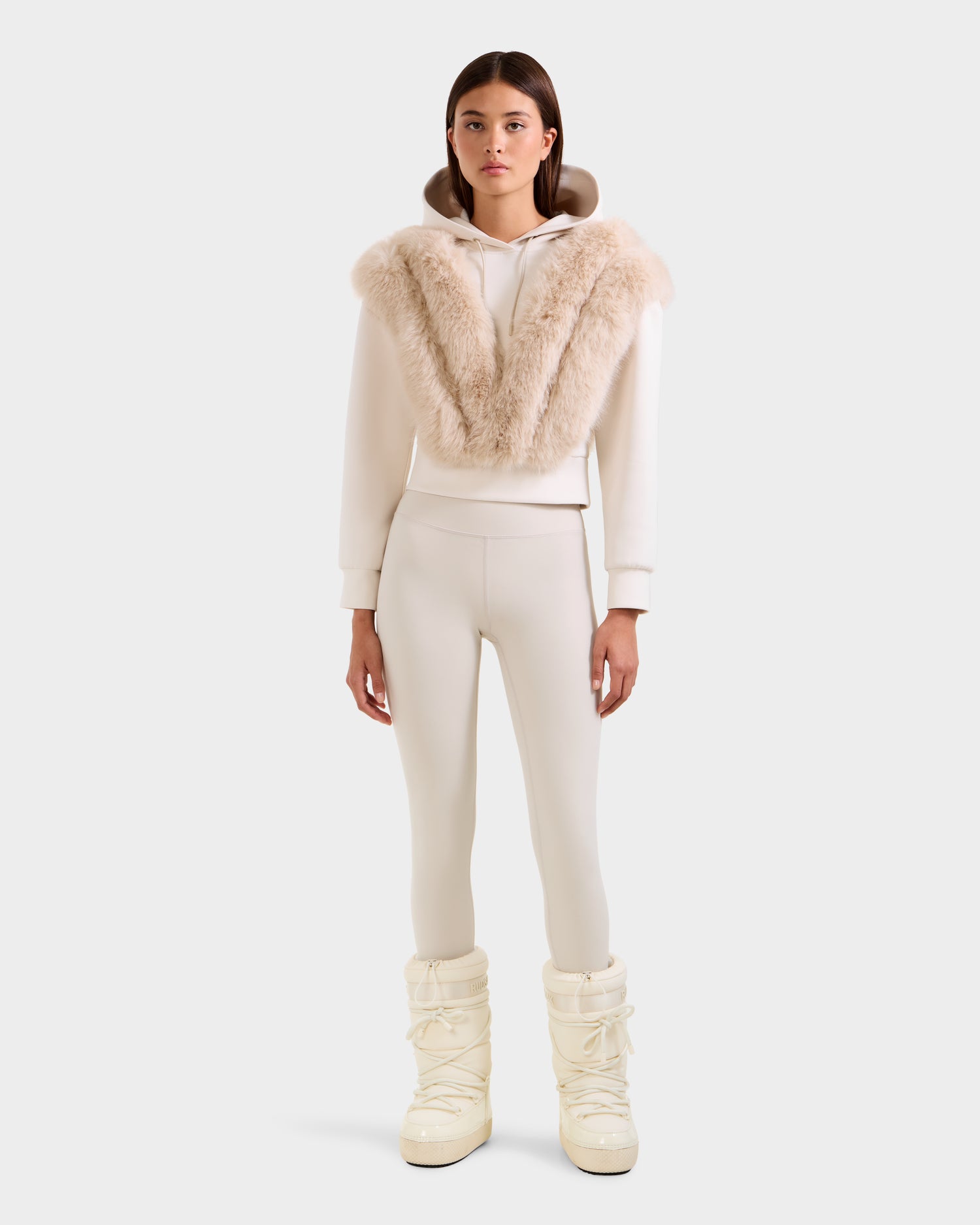 A woman faces forward in a cream-colored Rudsak DELLA - 8525668 Vanilla faux fur hoodie with a large collar, matching fitted pants, and white high winter boots against a plain light background.