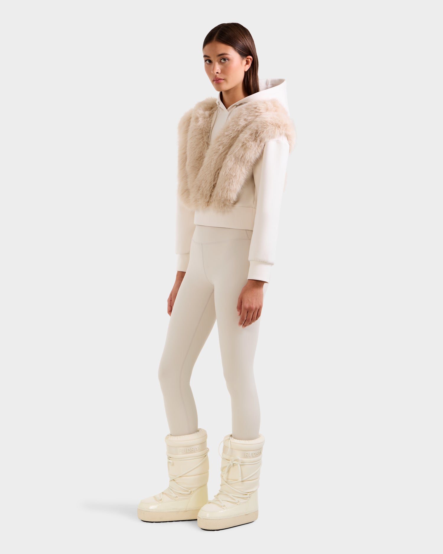 A woman models the Rudsak DELLA - 8525668 Vanilla ivory cropped women’s hoodie, paired with a faux fur vest, cream leggings, and off-white snow boots. She poses against a plain light background with straight brown hair.