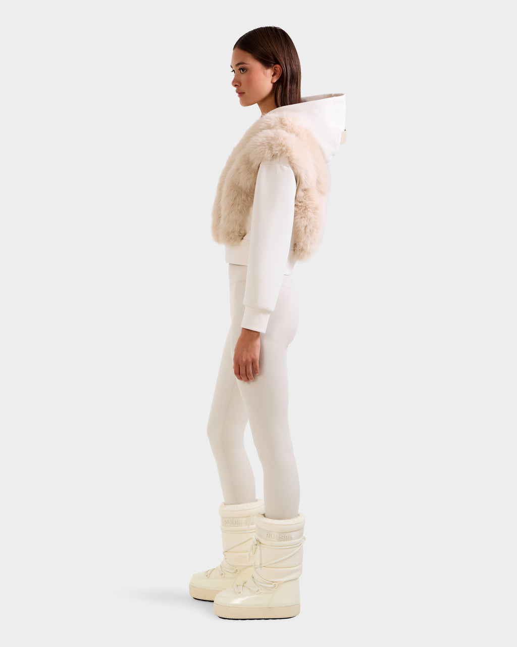 A woman stands in profile wearing the Rudsak DELLA - 8525668 Vanilla faux fur hoodie with a white pullover, white leggings, and off-white winter boots against a plain light background.
