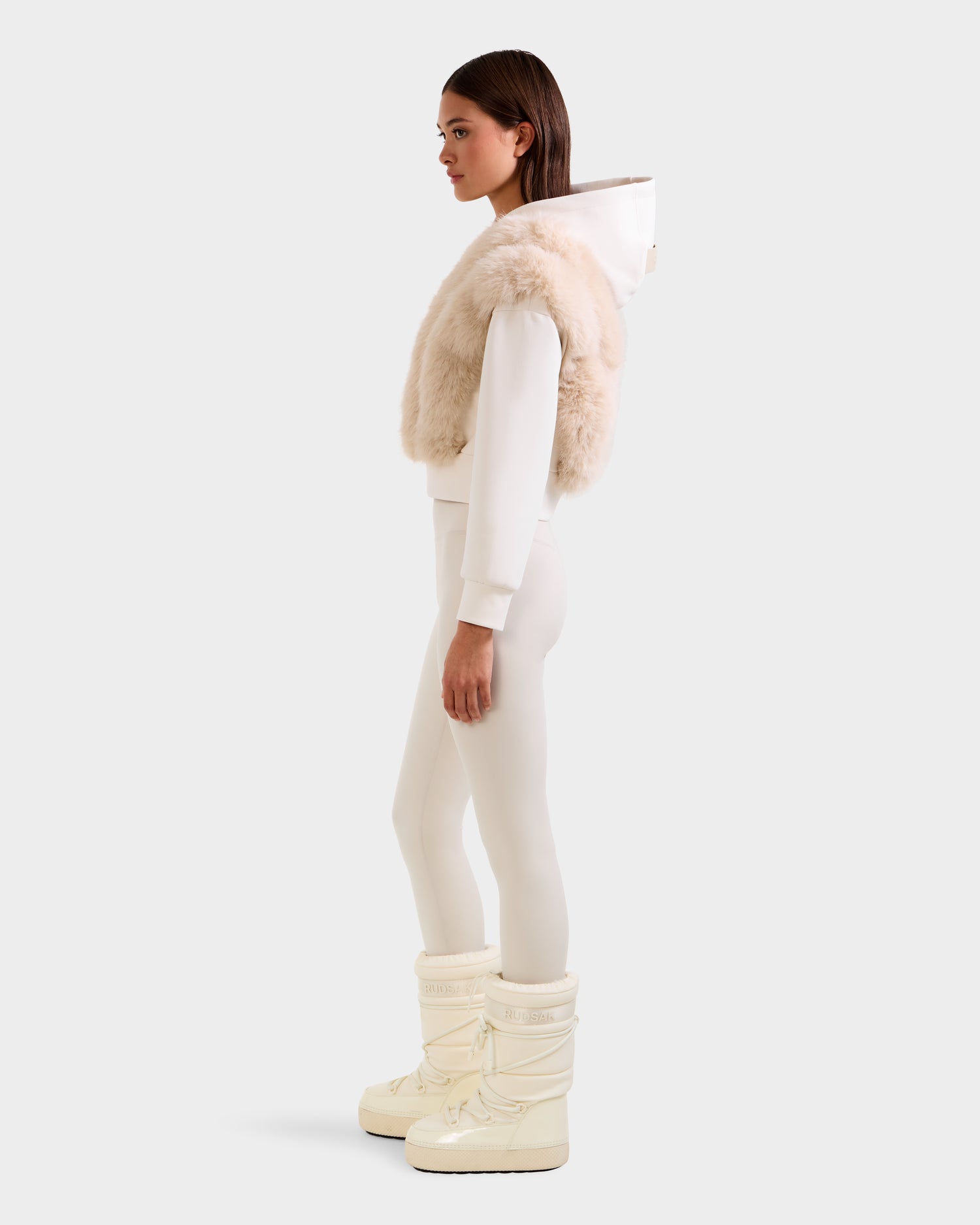 A woman stands in profile wearing the Rudsak DELLA - 8525668 Vanilla faux fur hoodie with a white pullover, white leggings, and off-white winter boots against a plain light background.
