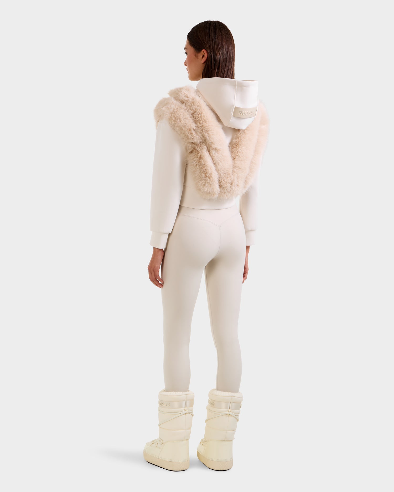 A woman stands facing away, wearing the Rudsak DELLA - 8525668 Vanilla faux fur hoodie with a beige fur shawl, off-white leggings, and cream snow boots against a plain light background.