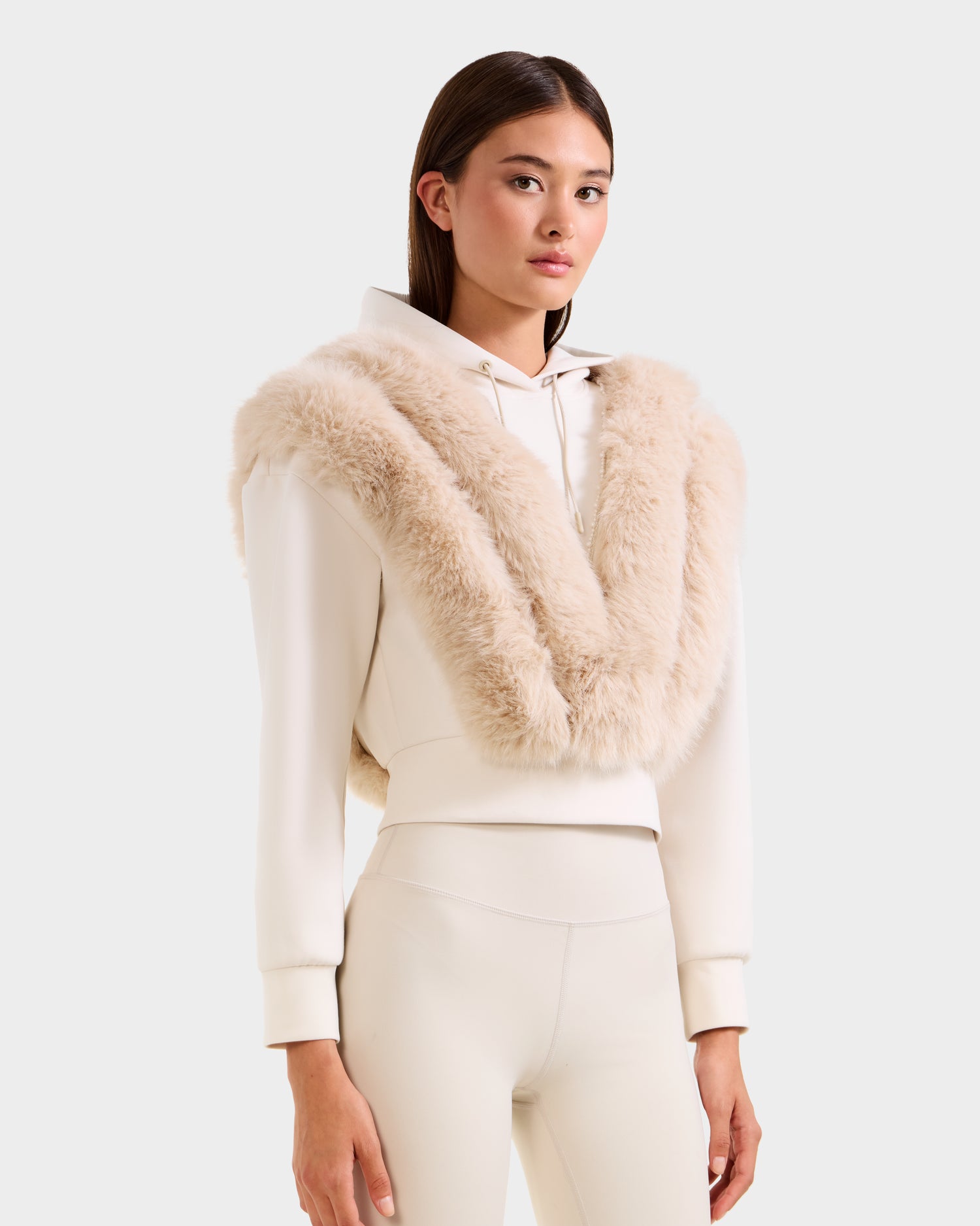 A woman faces forward in a Rudsak DELLA - 8525668 Vanilla après-ski pullover, featuring a wide faux fur shawl and matching fitted pants against a plain light gray background.