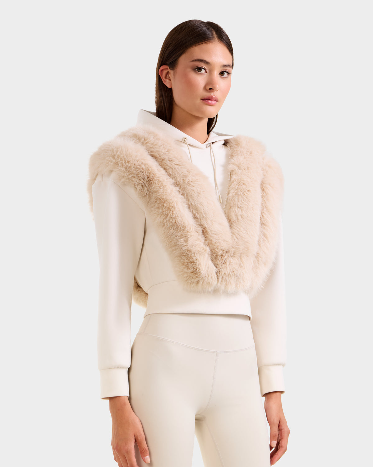 A woman faces forward in a Rudsak DELLA - 8525668 Vanilla après-ski pullover, featuring a wide faux fur shawl and matching fitted pants against a plain light gray background.