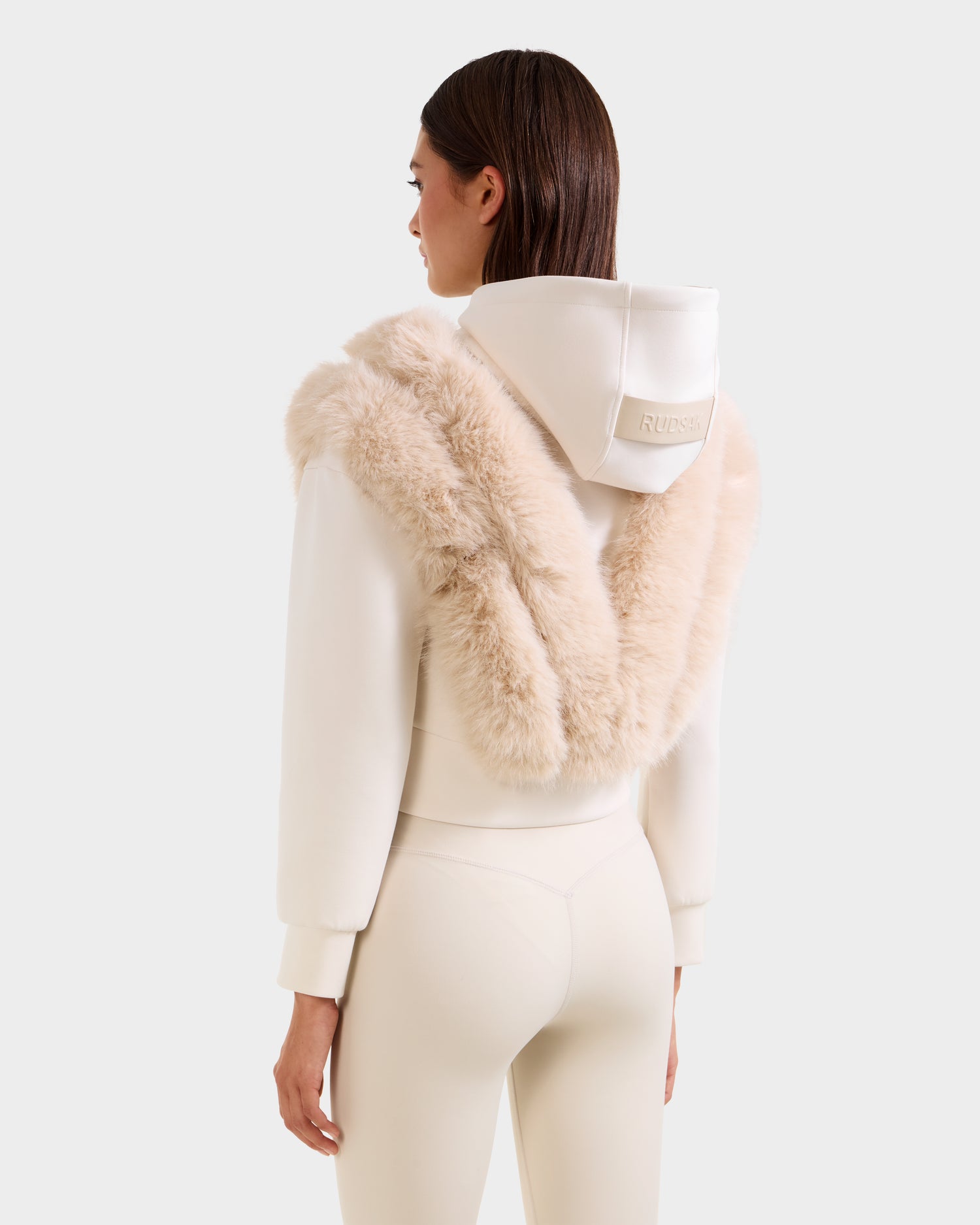 A woman faces away, wearing the Rudsak DELLA - 8525668 Vanilla faux fur hoodie with plush trim over the hood and shoulders, paired with matching leggings—a chic après-ski pullover look.