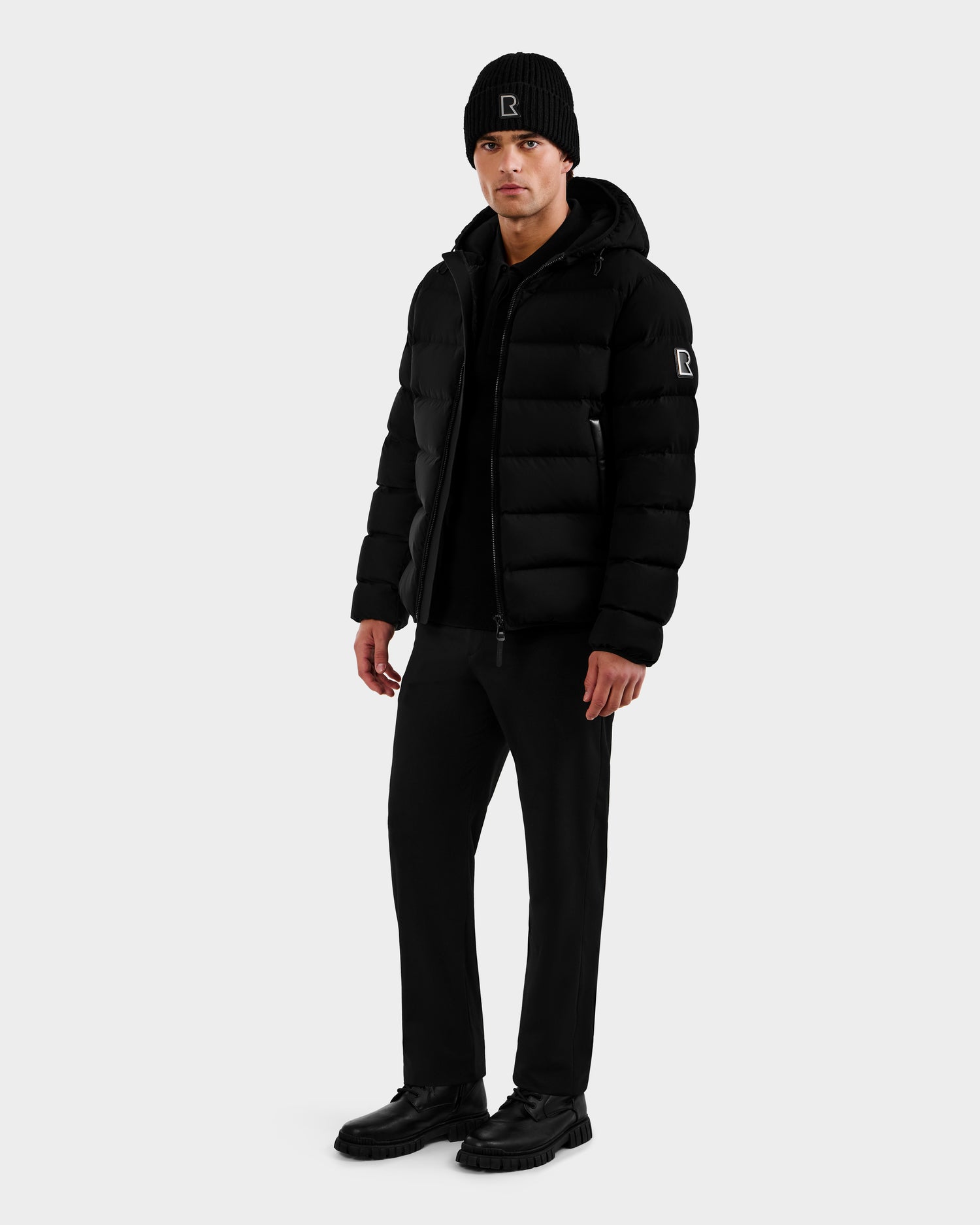 A man in Rudsak’s DEMIAN M - M123507 Black down puffer stands slightly turned to the side, pairing the jacket with a black beanie, pants, and shoes against a plain light background.
