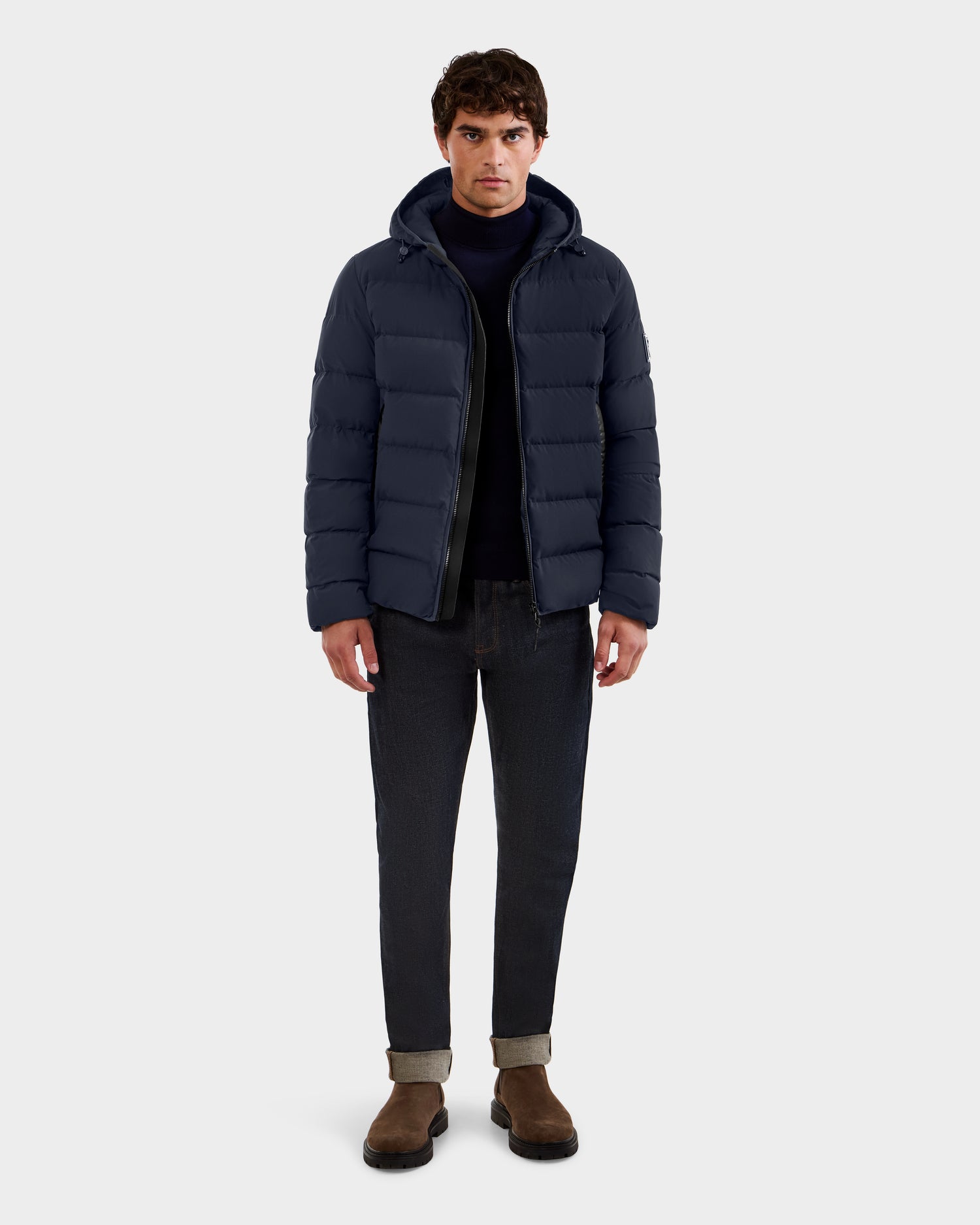 A man faces forward wearing the Rudsak DEMIAN M - M123507 Navy down puffer jacket, crafted from sustainable recycled fabric, styled with a dark sweater, cuffed black jeans, and brown suede boots against a light gray background.