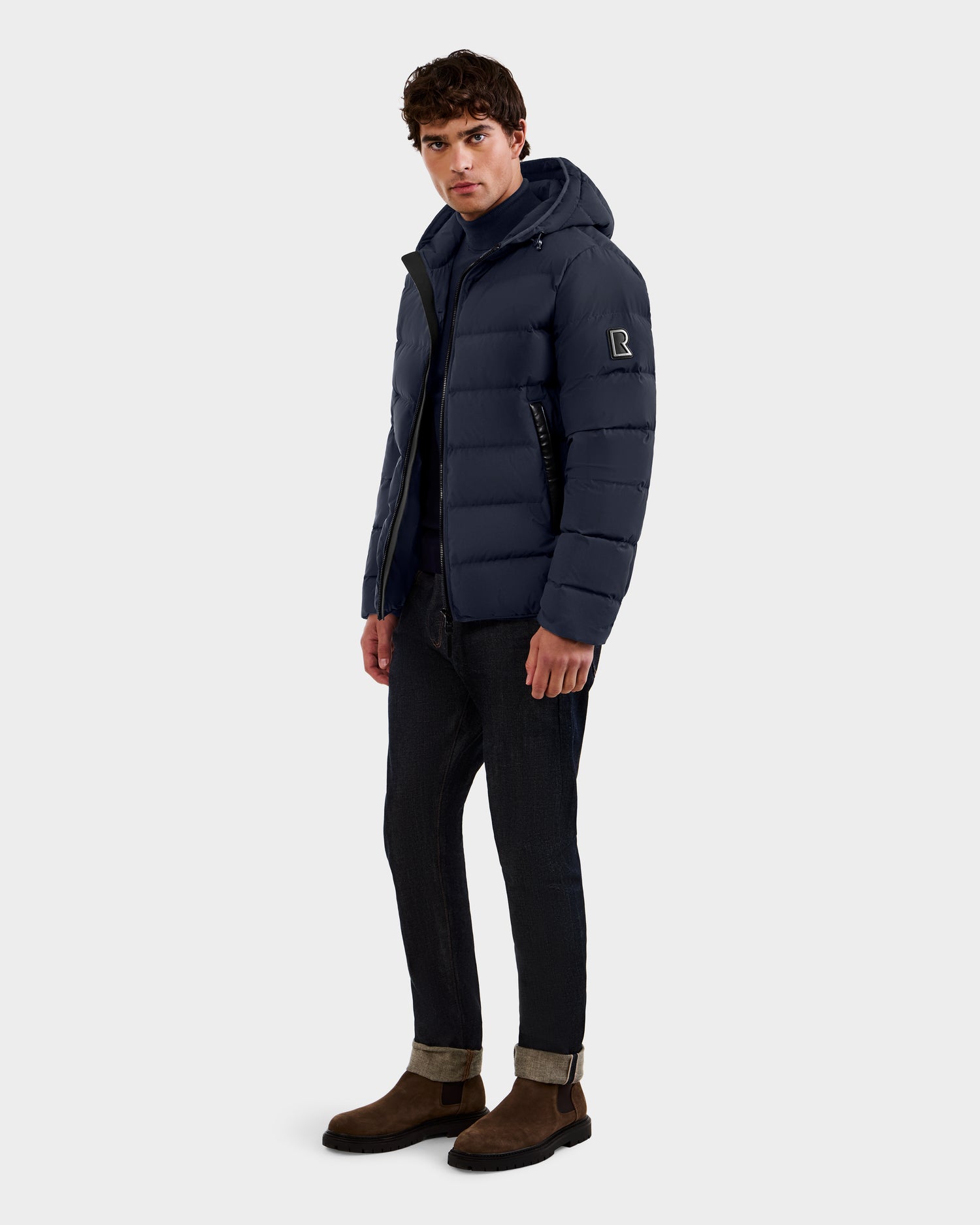 A man stands facing forward in the Rudsak DEMIAN M - M123507 Navy men’s down puffer jacket, made from RDS-certified premium down and recycled fabric. He pairs it with dark cuffed jeans and brown suede boots against a plain light gray background.