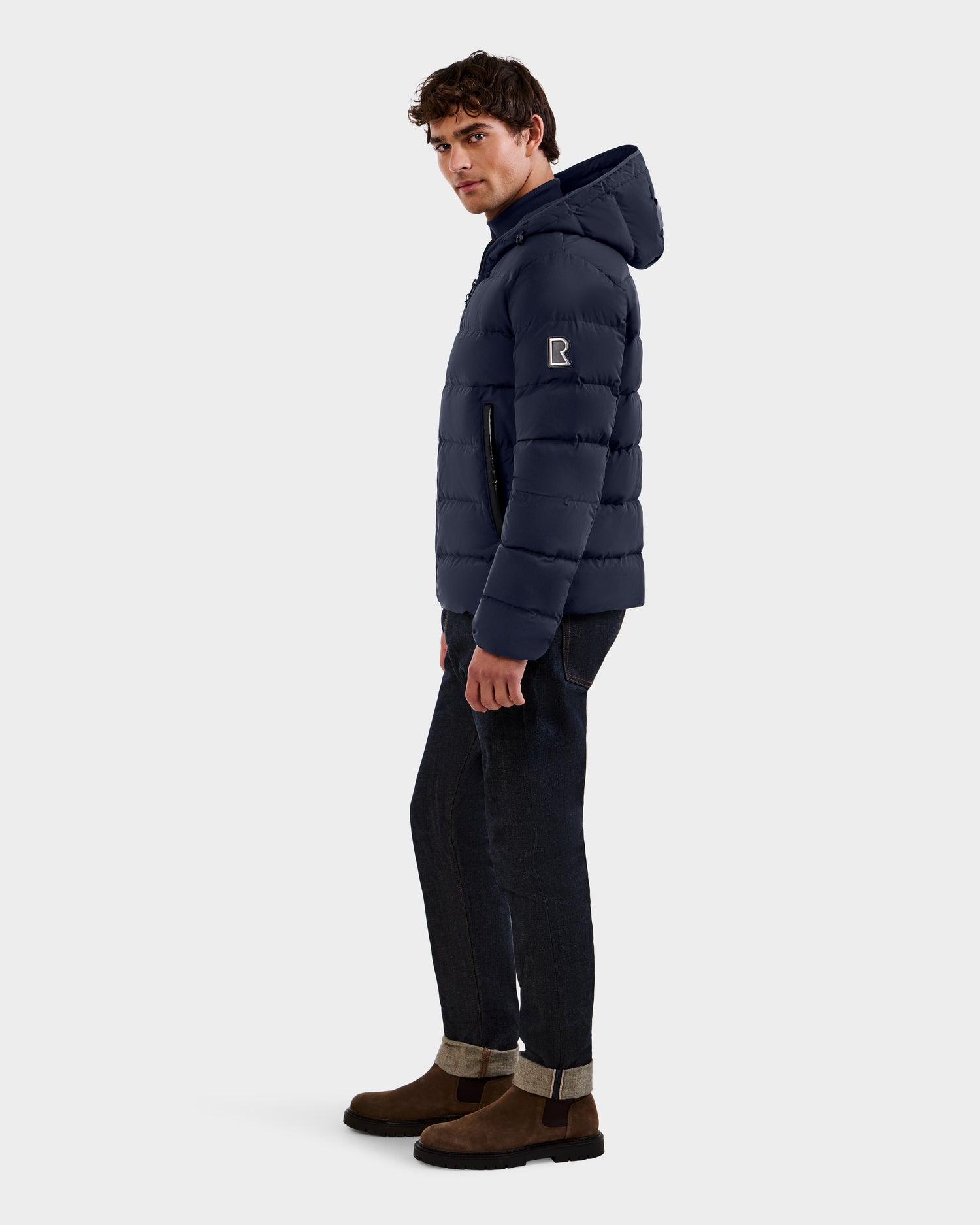 A man in the Rudsak DEMIAN M - M123507 Navy puffer jacket made from sustainable recycled fabric, with dark jeans and brown boots, stands sideways and looks over his shoulder against a plain white background.