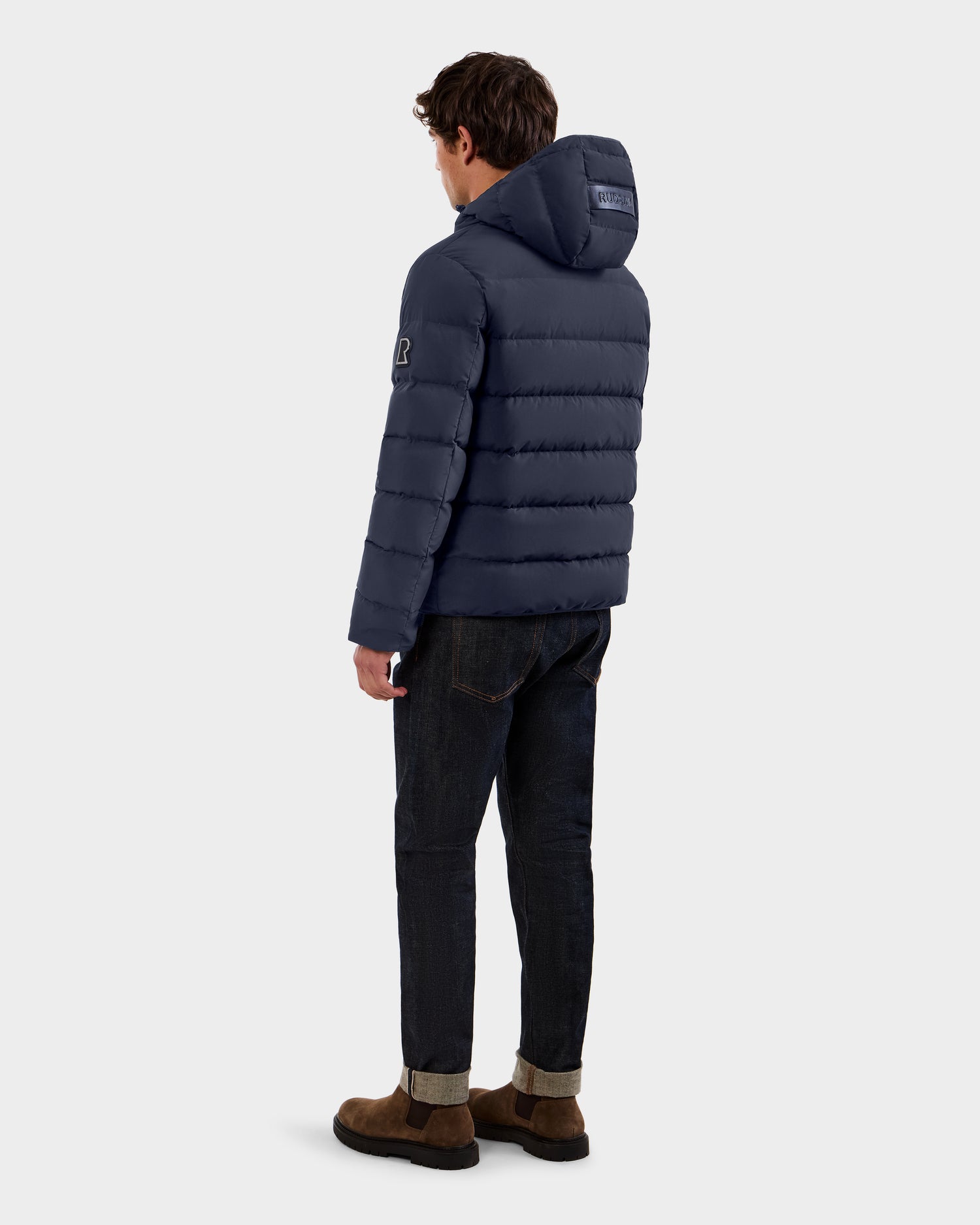 A man stands facing away, wearing the Rudsak DEMIAN M - M123507 navy hooded down puffer made from sustainable recycled fabric, paired with dark jeans and brown suede shoes against a plain light background.