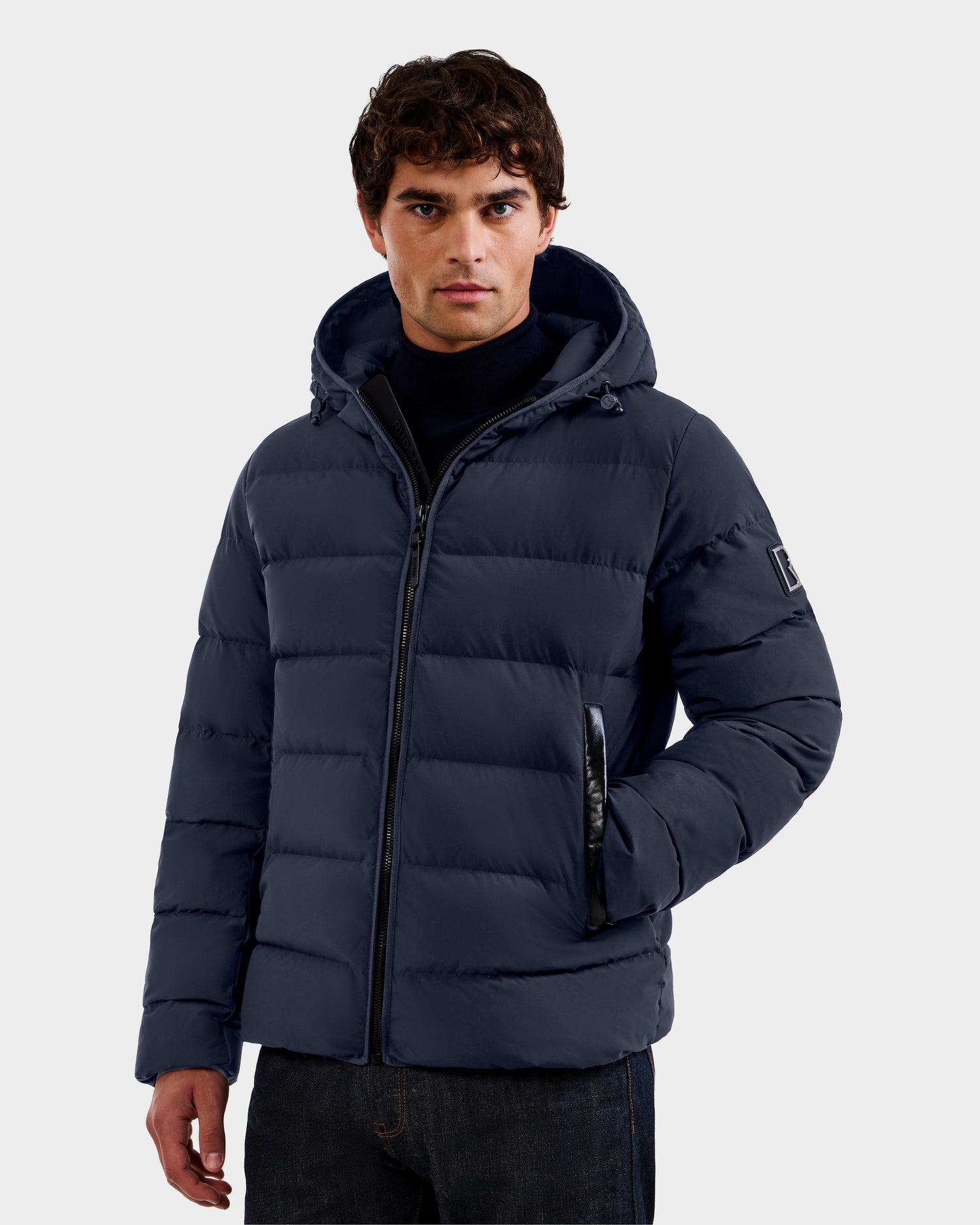 A man wearing the Rudsak DEMIAN M - M123507 Navy hooded down puffer made from sustainable recycled fabric stands against a plain light background, with one hand in his pocket and a neutral expression, looking at the camera.