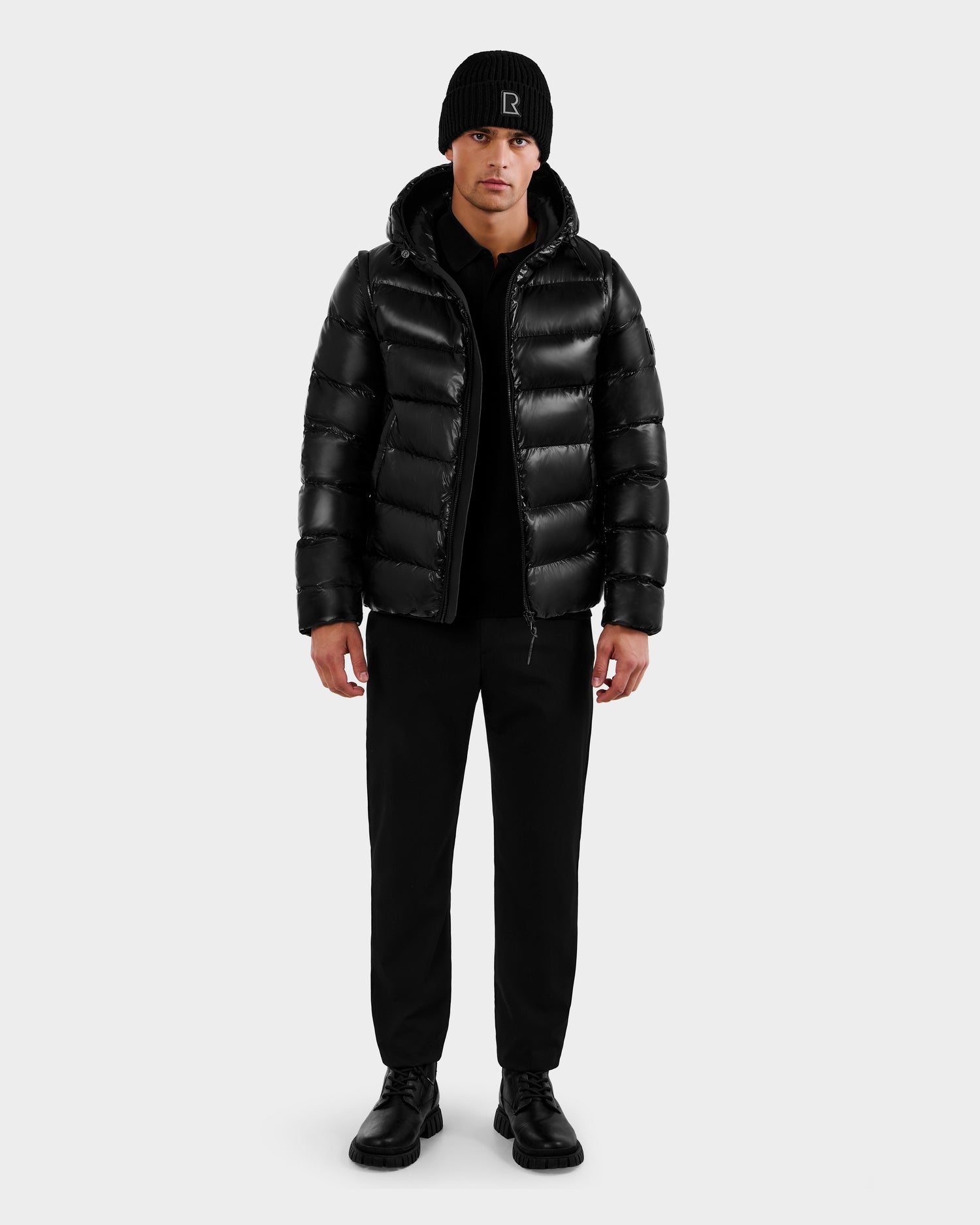 A man faces forward wearing the Rudsak DEMIAN S-R - M123505 Black down puffer jacket, paired with black pants, shoes, and a beanie with a small logo. The background is plain and light-colored.