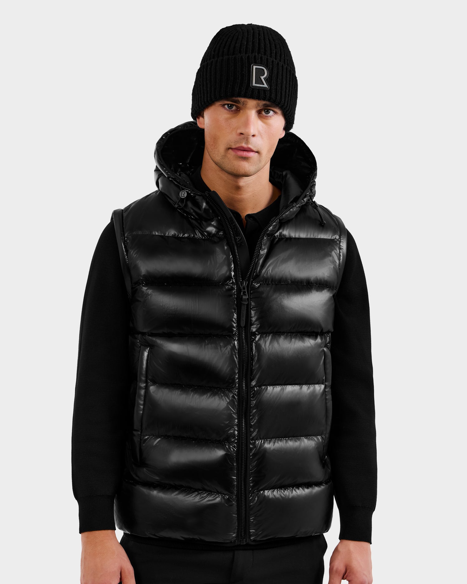 A man in a Rudsak DEMIAN S-R - M123505 Black 2-in-1 puffer jacket with hood, black knit beanie, and black long-sleeve shirt stands facing forward against a plain light gray background.