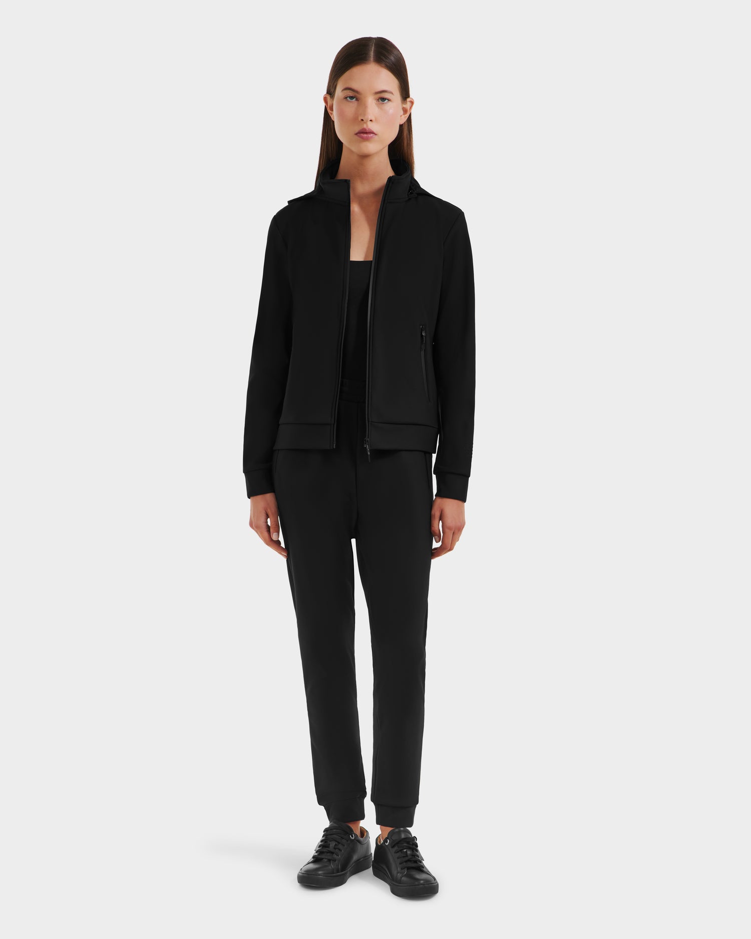A woman with straight brown hair wears the black DERIAM - 8525573 zip-up sweater by Rudsak, paired with matching black pants and sneakers. She faces forward with a neutral expression against a plain light background.