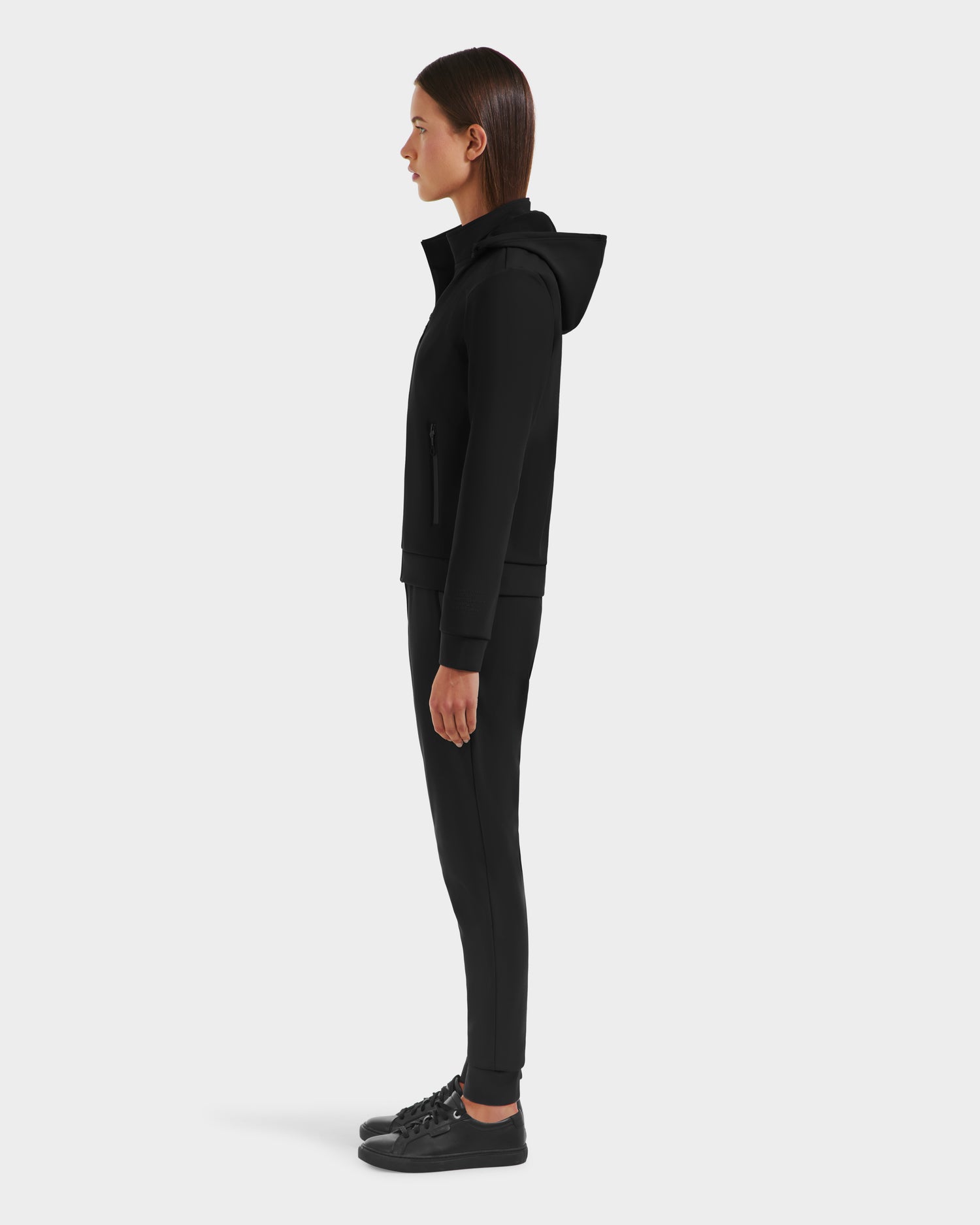 A person with straight brown hair stands in profile, facing left, wearing the Rudsak DERIAM - 8525573 Black women’s sporty zip-up sweater with removable hood, along with black pants and sneakers, against a plain white background.