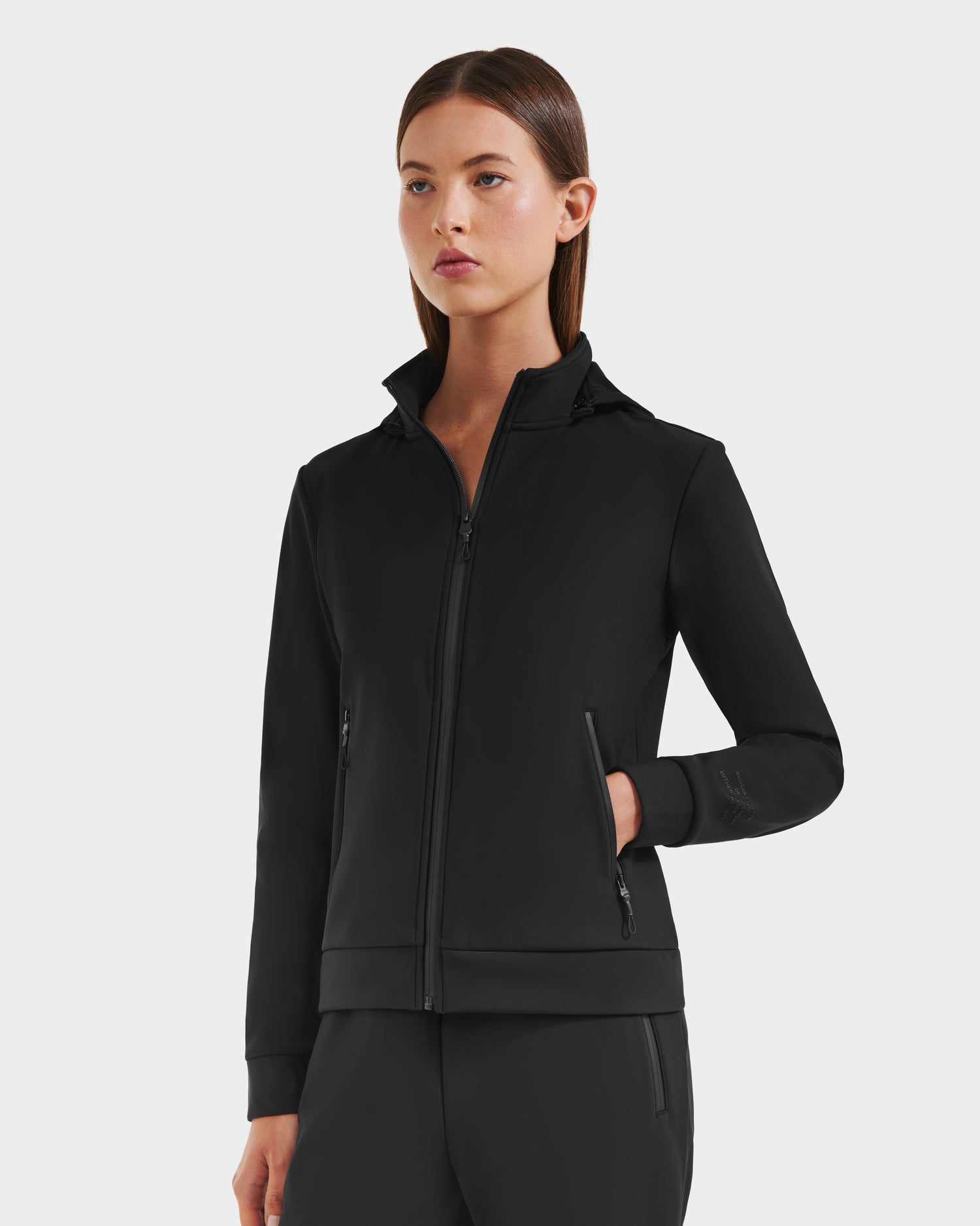 A woman with straight brown hair wears the Rudsak DERIAM - 8525573 Black zip-up sweater jacket and matching pants, made from performance stretch fabric, standing against a light gray background with one hand in her pocket.