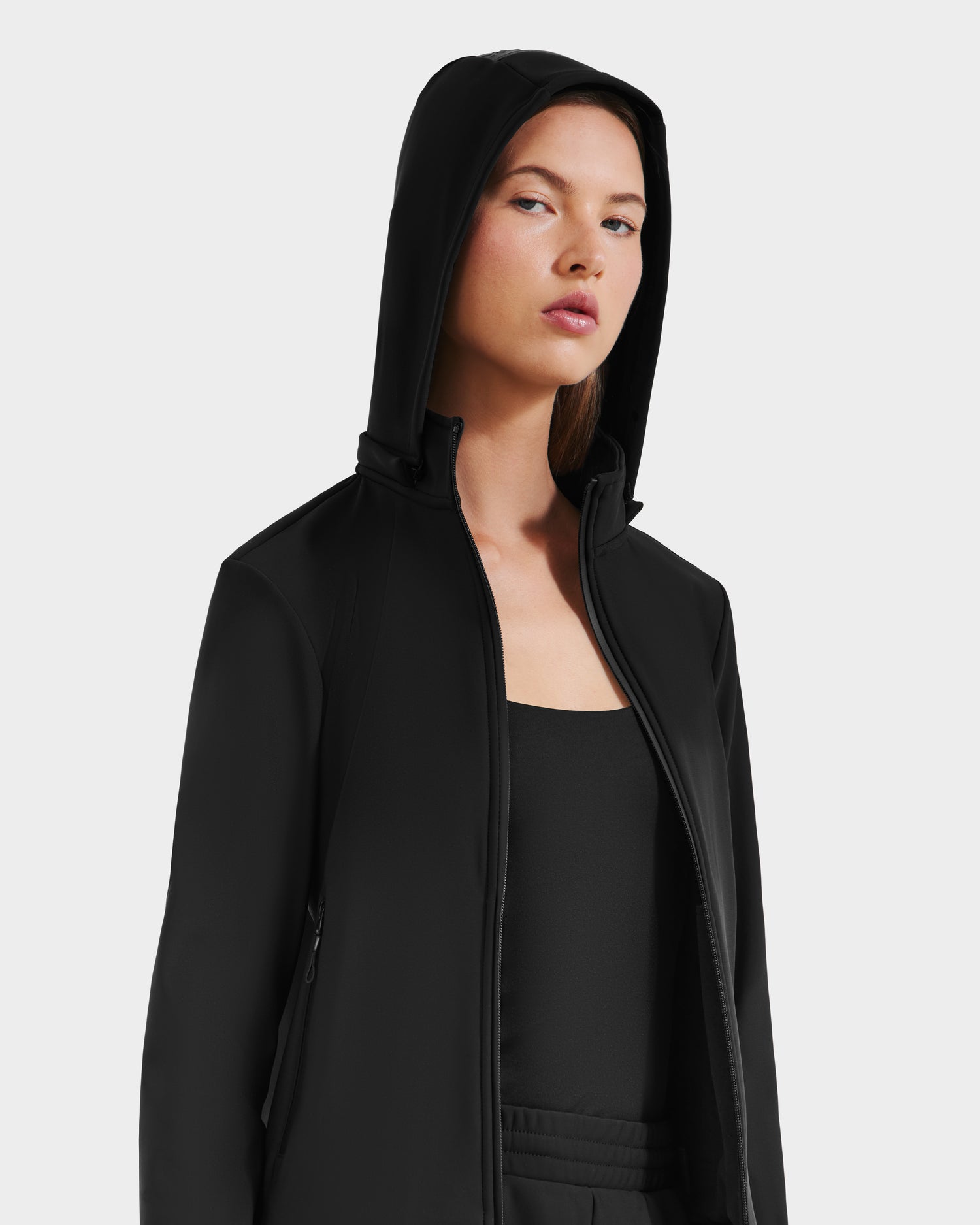 A woman wears the black DERIAM - 8525573 zip-up sweater by Rudsak over a black top, hood up and unzipped, against a plain light backdrop, highlighting her sporty style and neutral expression.