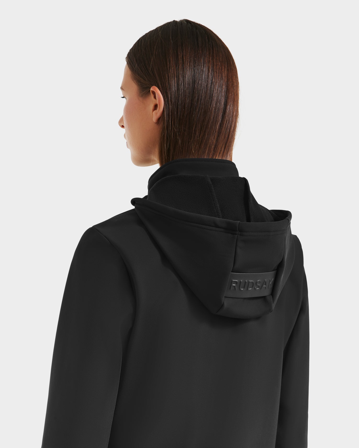 A woman with straight brown hair, seen from behind, wears the Rudsak DERIAM - 8525573 Black jacket with a logo patch on the back of its removable hood. The background is plain and light-colored.