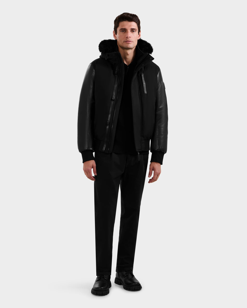 A man faces forward in the Rudsak DERON - M125539 Black down bomber with vegan-leather sleeves, paired with black pants and shoes, against a plain light background.