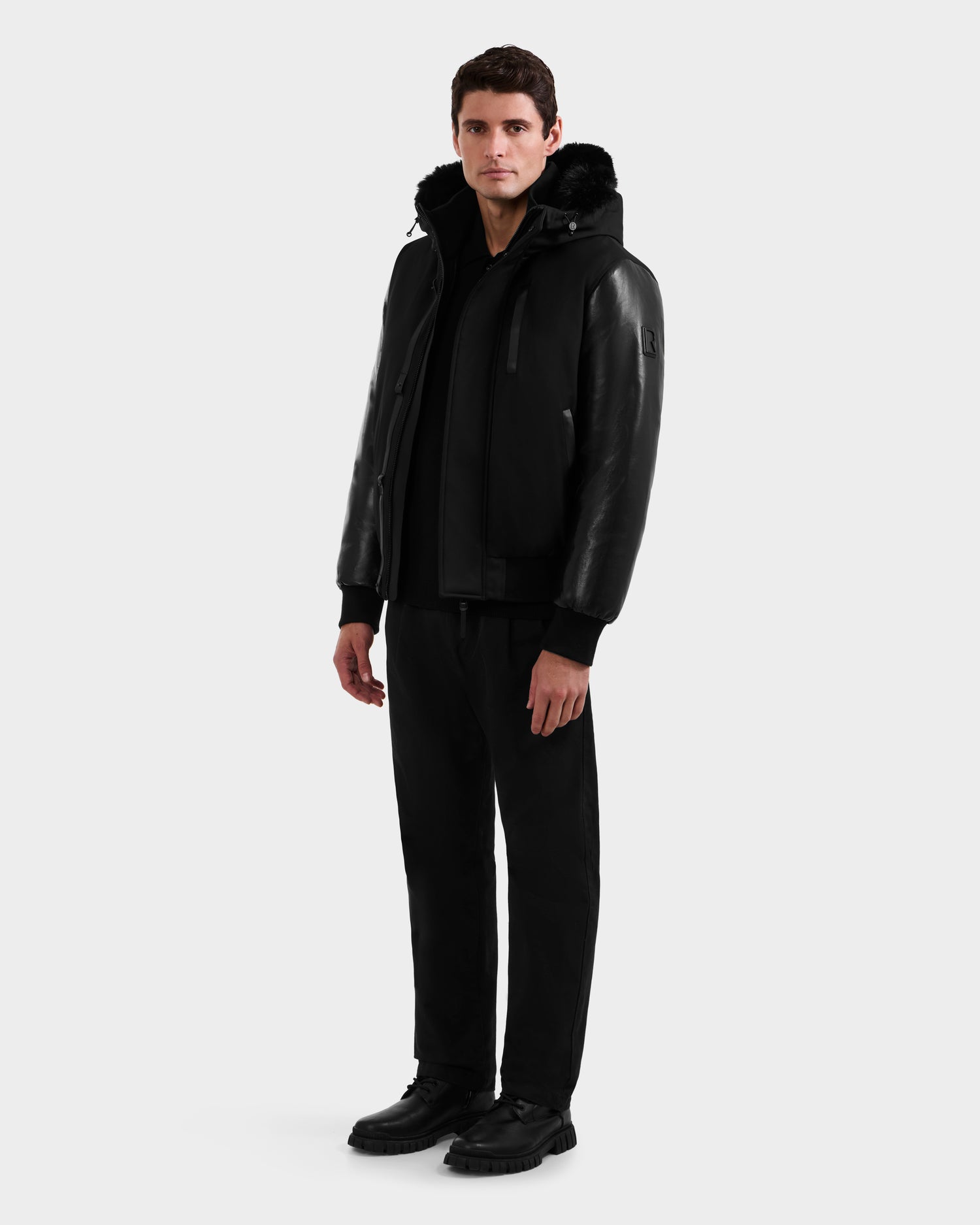 A man faces forward in a Rudsak DERON - M125539 Black jacket with vegan-leather sleeves, black pants, and black shoes against a plain white background.