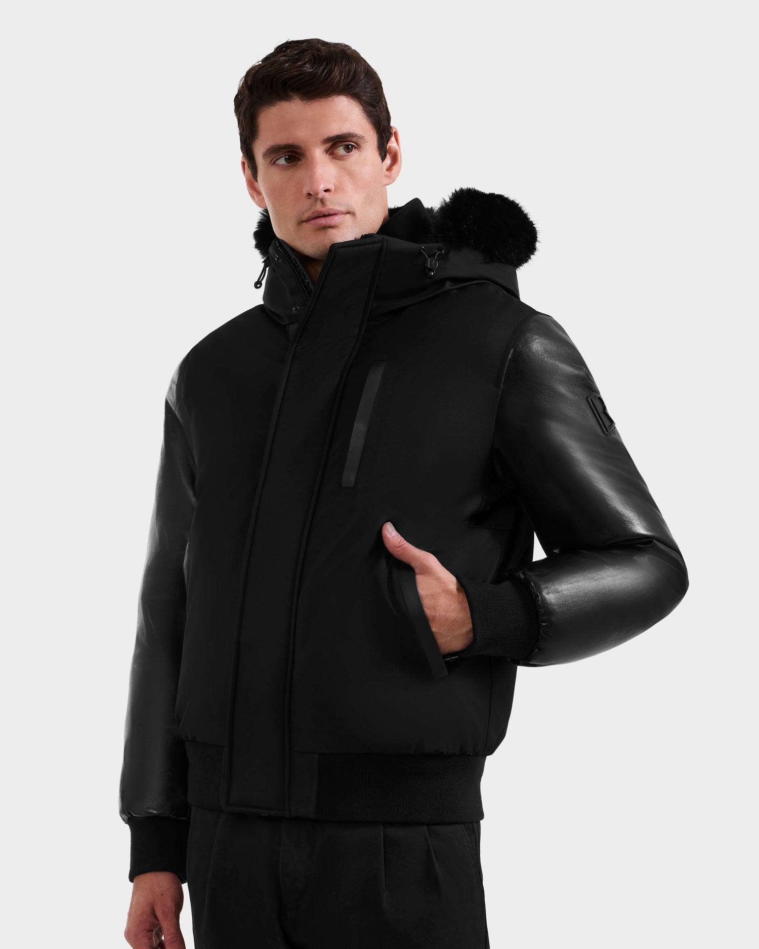 A man stands against a plain white background wearing the Rudsak DERON - M125539 Black down bomber with vegan-leather sleeves and a fur-trimmed hood, looking slightly to the side with one hand in his pocket.