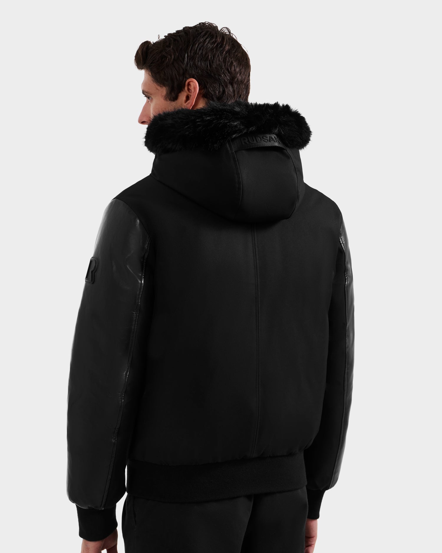 A man is seen from behind wearing the Rudsak DERON - M125539 Black men’s down bomber with vegan-leather sleeves, faux fur trim, and glossy accents against a plain light gray background.