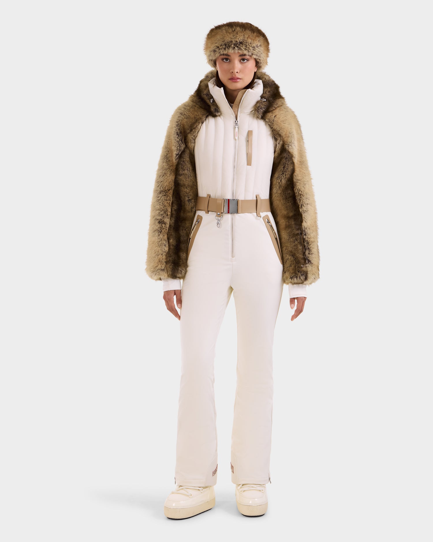 A woman faces forward wearing the Rudsak DESTINY - 8124727 Vanilla women's ski suit with beige accents, a belted waist, brown faux fur jacket, matching fur hat, and cream platform boots—set against a plain white background.