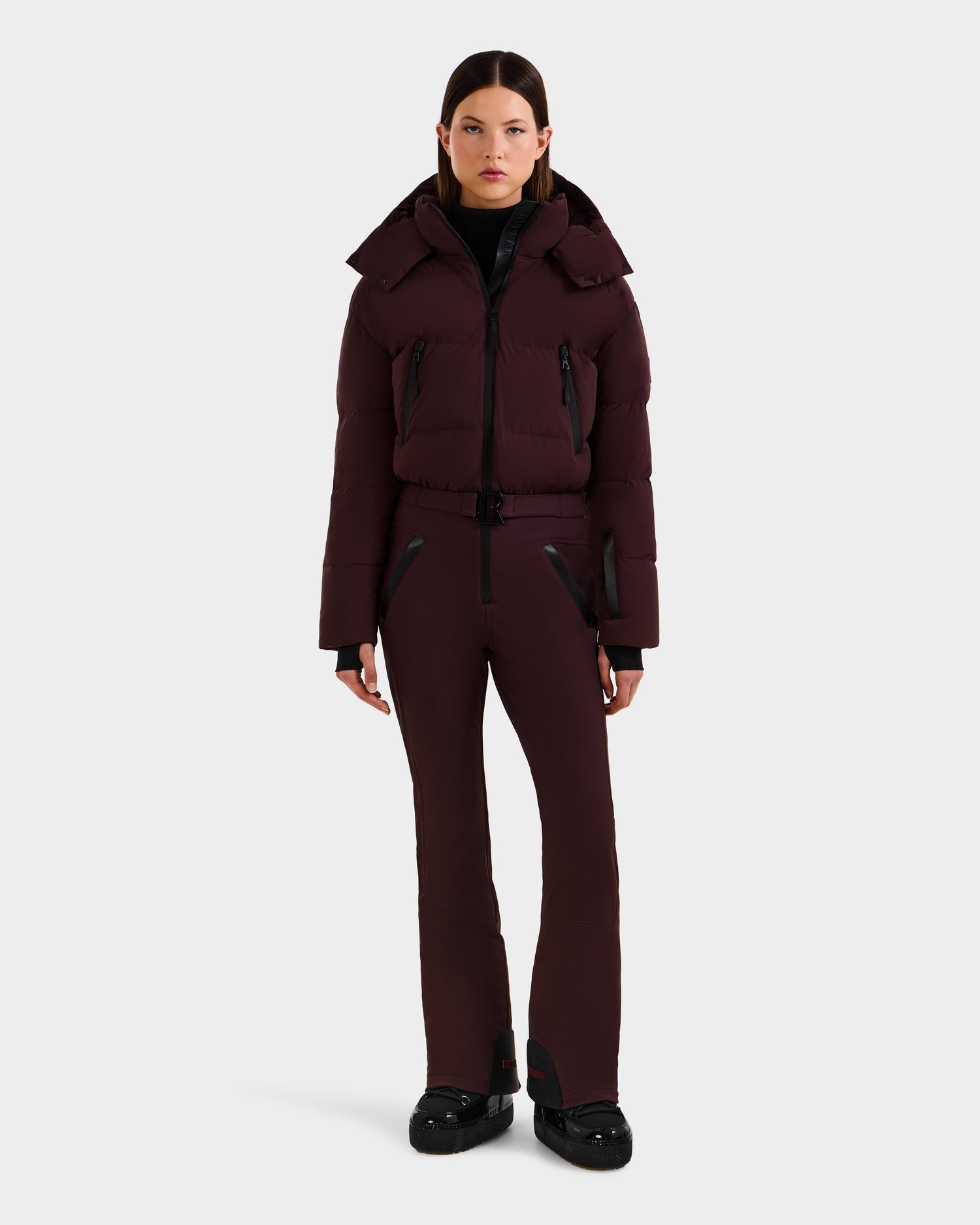 A woman stands against a plain background wearing the Rudsak DINARA - 8125600 Bordeaux women’s ski suit with black details and black shoes, facing forward with relaxed arms—a chic après-ski look.