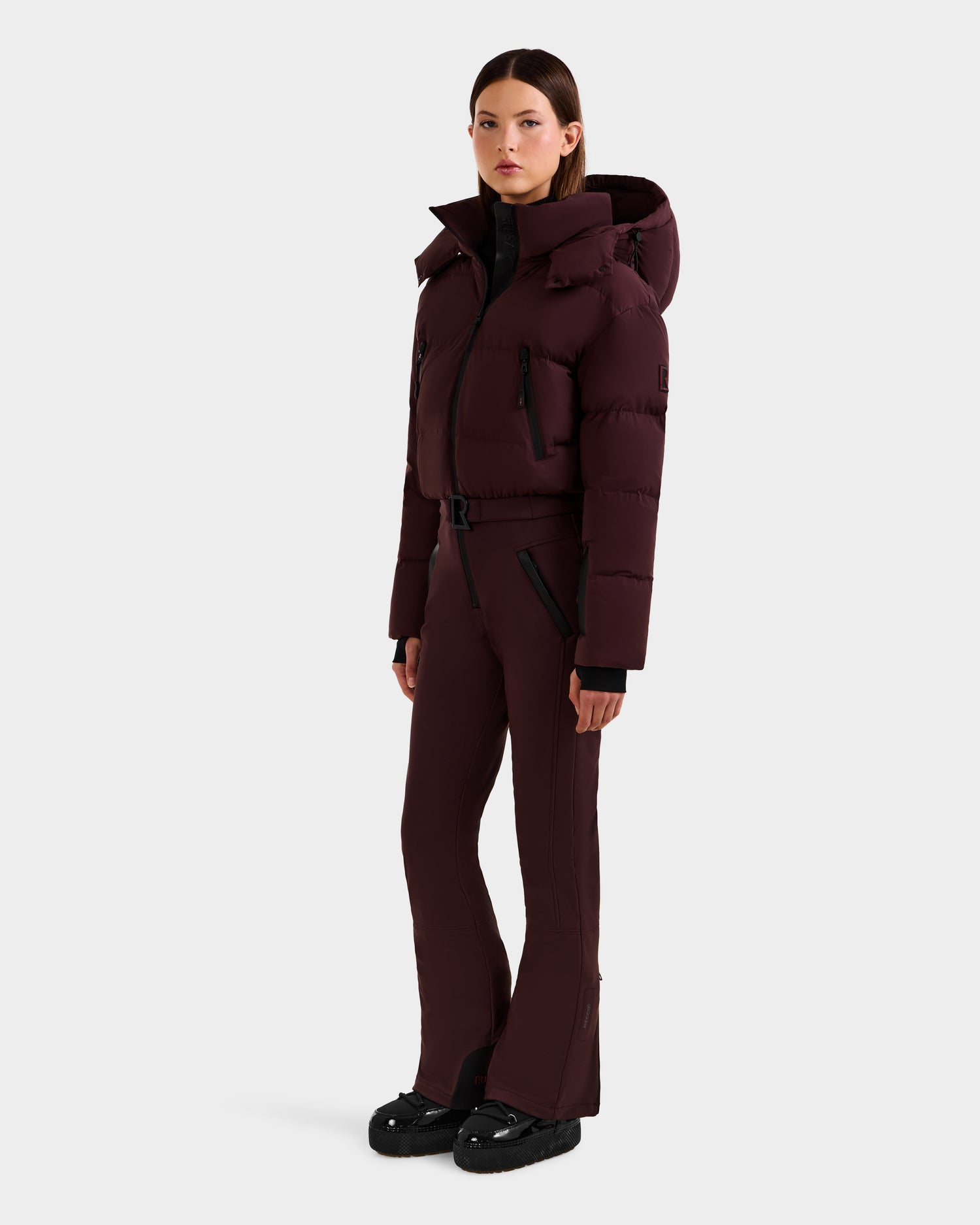 A woman models the Rudsak DINARA - 8125600 Bordeaux ski suit: a hooded maroon puffer jacket and matching fitted pants with black zippers, paired with black boots, styled for après-ski, set against a plain white background.