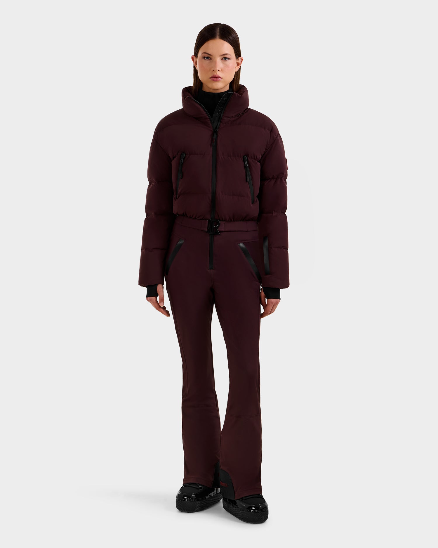A woman faces forward in the Rudsak DINARA - 8125600 Bordeaux ski suit, featuring a sleek dark burgundy puffer jacket with black accents and matching fitted pants, paired with black shoes against a plain, light background.
