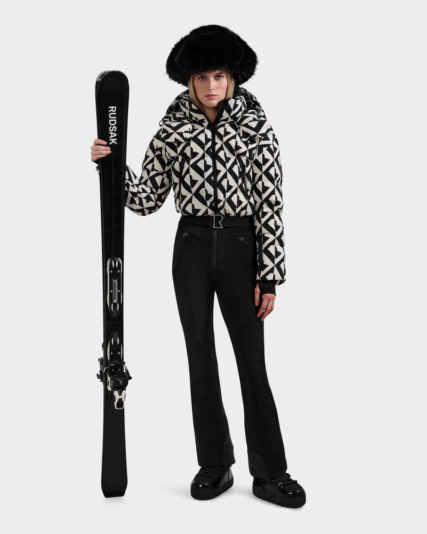 A woman wearing the Rudsak DINARA - 8125600 Vanilla Monogram ski suit and black boots, with a large black fur hat, stands holding black skis upright against a plain background.