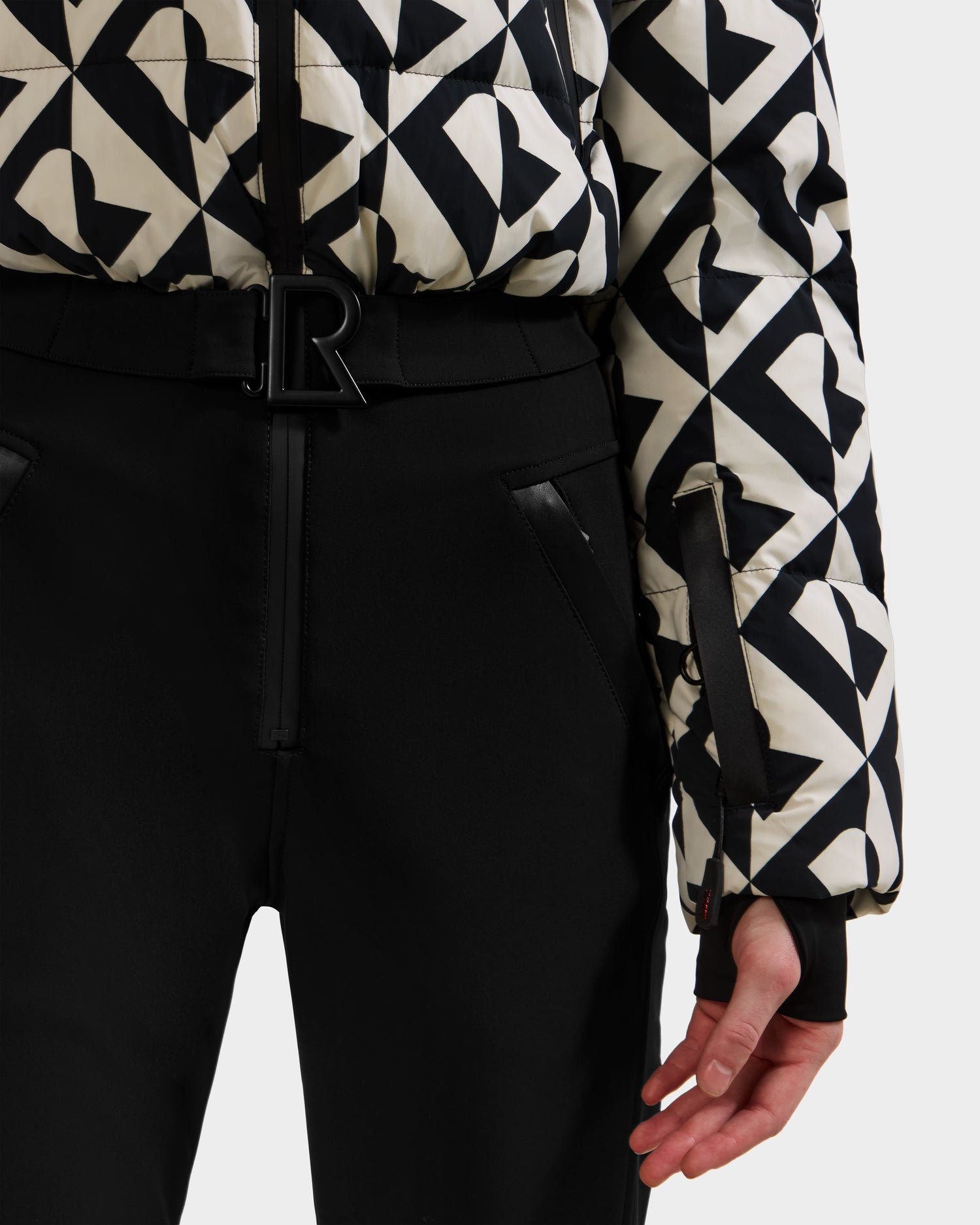 A person wearing black pants and a Rudsak DINARA - 8125600 Vanilla Monogram jacket with a black belt and large R buckle, resembling a chic ski suit. Only the lower torso and one hand are visible.