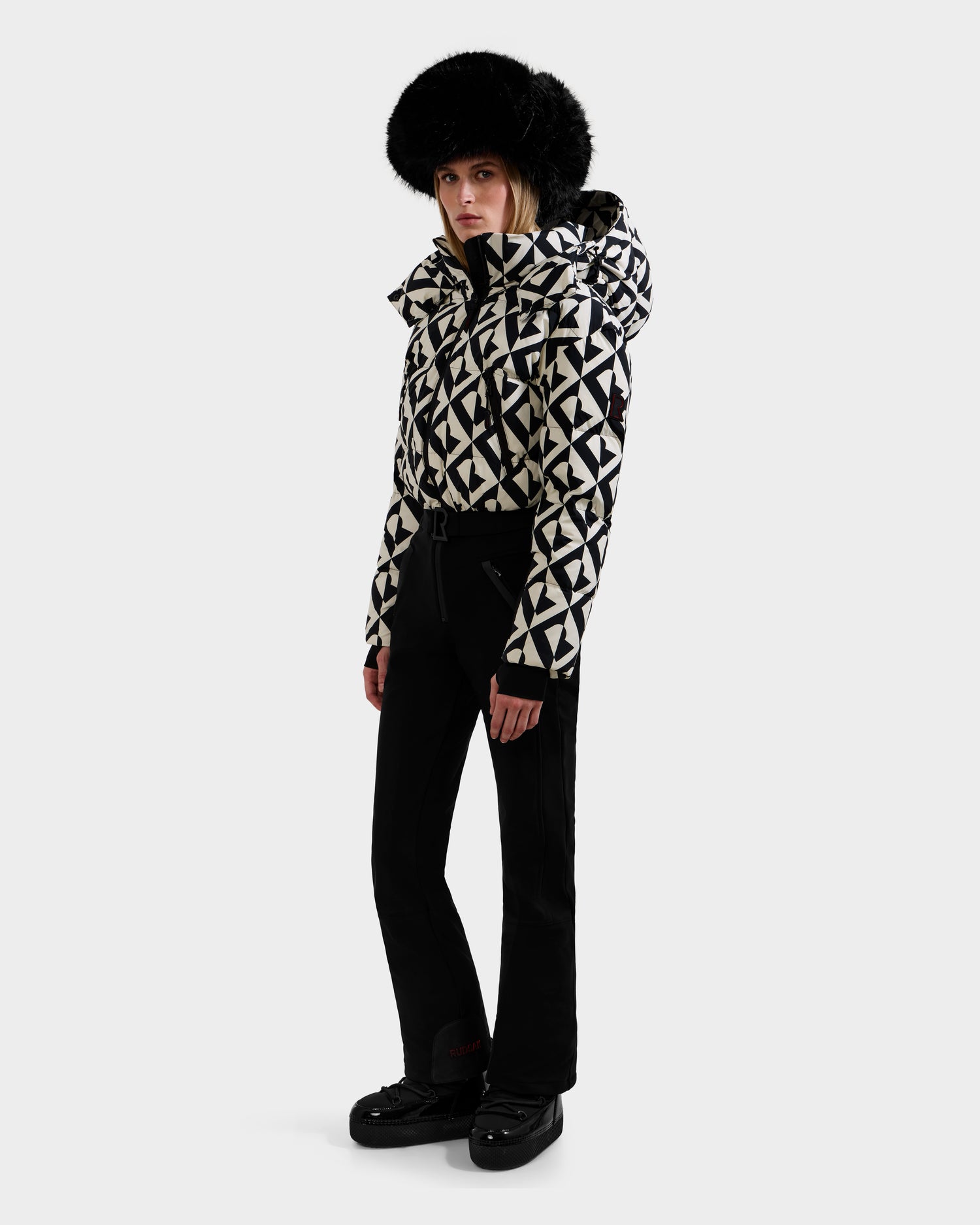 A person wears the Rudsak DINARA - 8125600 Vanilla Monogram women's ski suit and black boots with a black fur hat, standing against a plain white background.