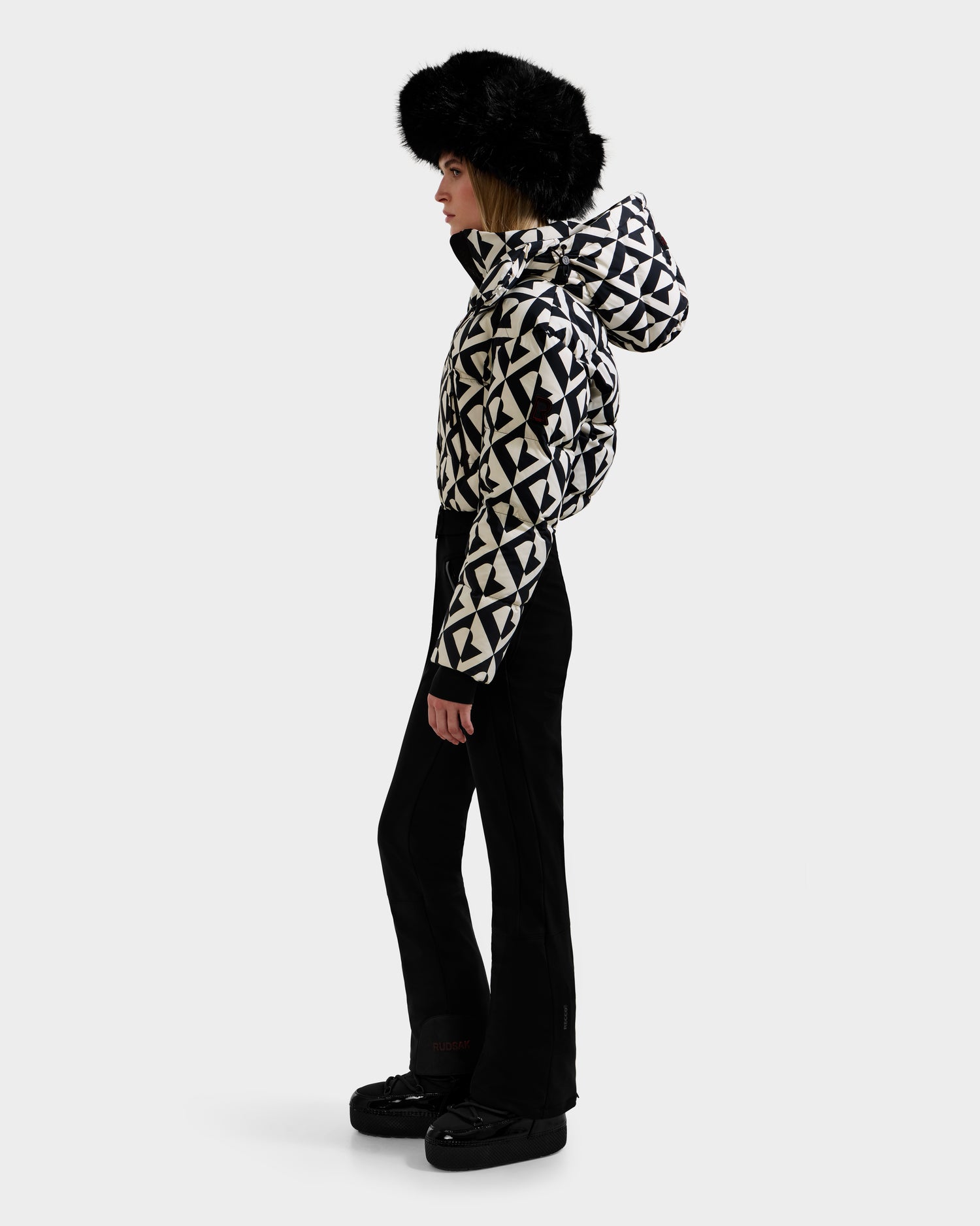 A person stands in profile against a white background wearing the Rudsak DINARA - 8125600 Vanilla Monogram women's ski suit with hood, black flared pants, black winter boots, and a large black fur hat.