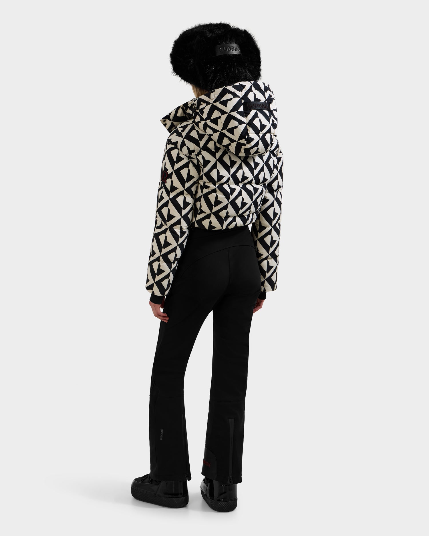 A person faces away, wearing black pants, boots, and the Rudsak DINARA - 8125600 Vanilla Monogram women’s ski suit—a white and black geometric-patterned jacket with a hood trimmed in black fur—against a plain white background.