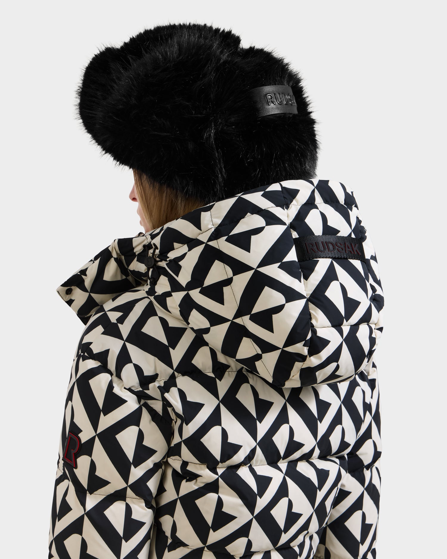 A woman faces away from the camera wearing Rudsak’s DINARA - 8125600 Vanilla Monogram puffer jacket, a women’s ski suit style with a black and white geometric pattern, large hood, RECCO rescue tech on the back, and a black fur hat.